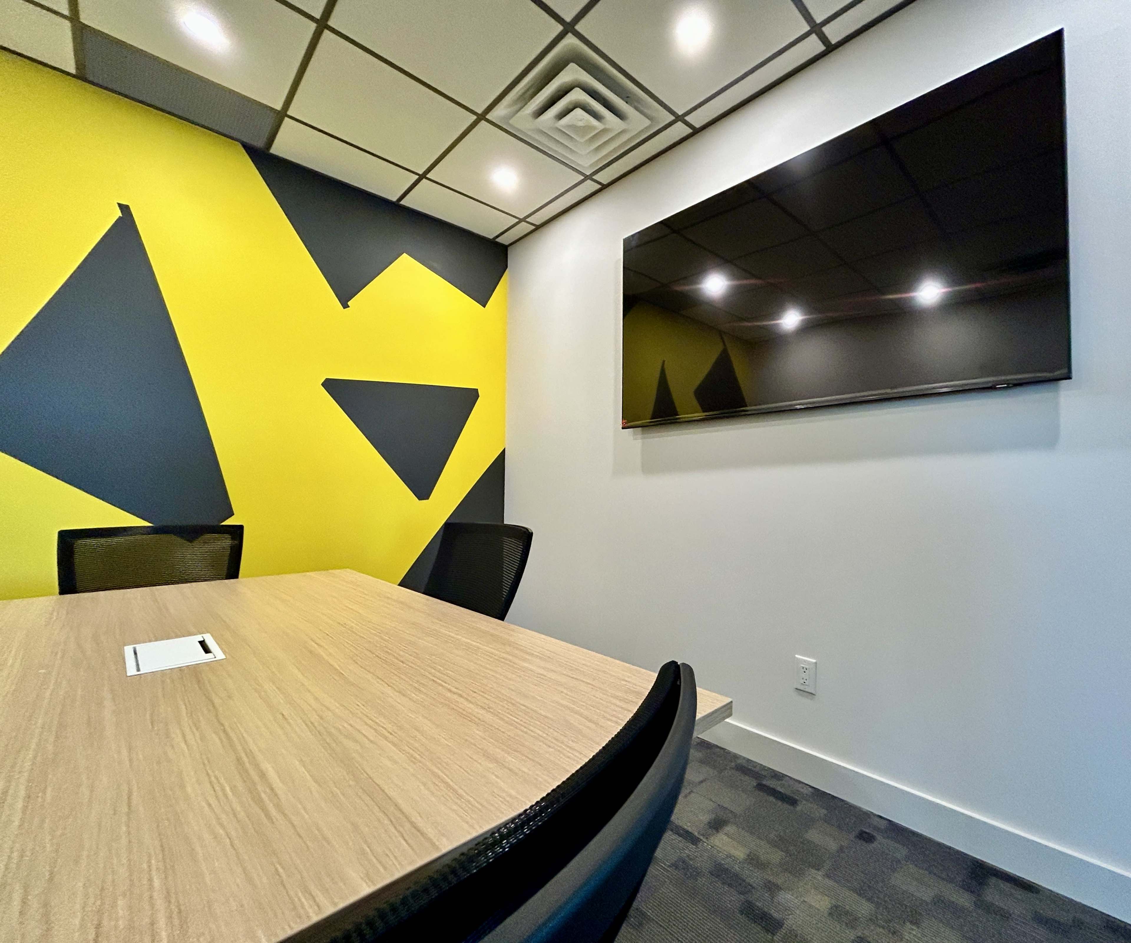 A modern conference room features a wooden table, black chairs, and a wall with a yellow and black geometric design, along with a flat-screen television mounted on the opposite wall.