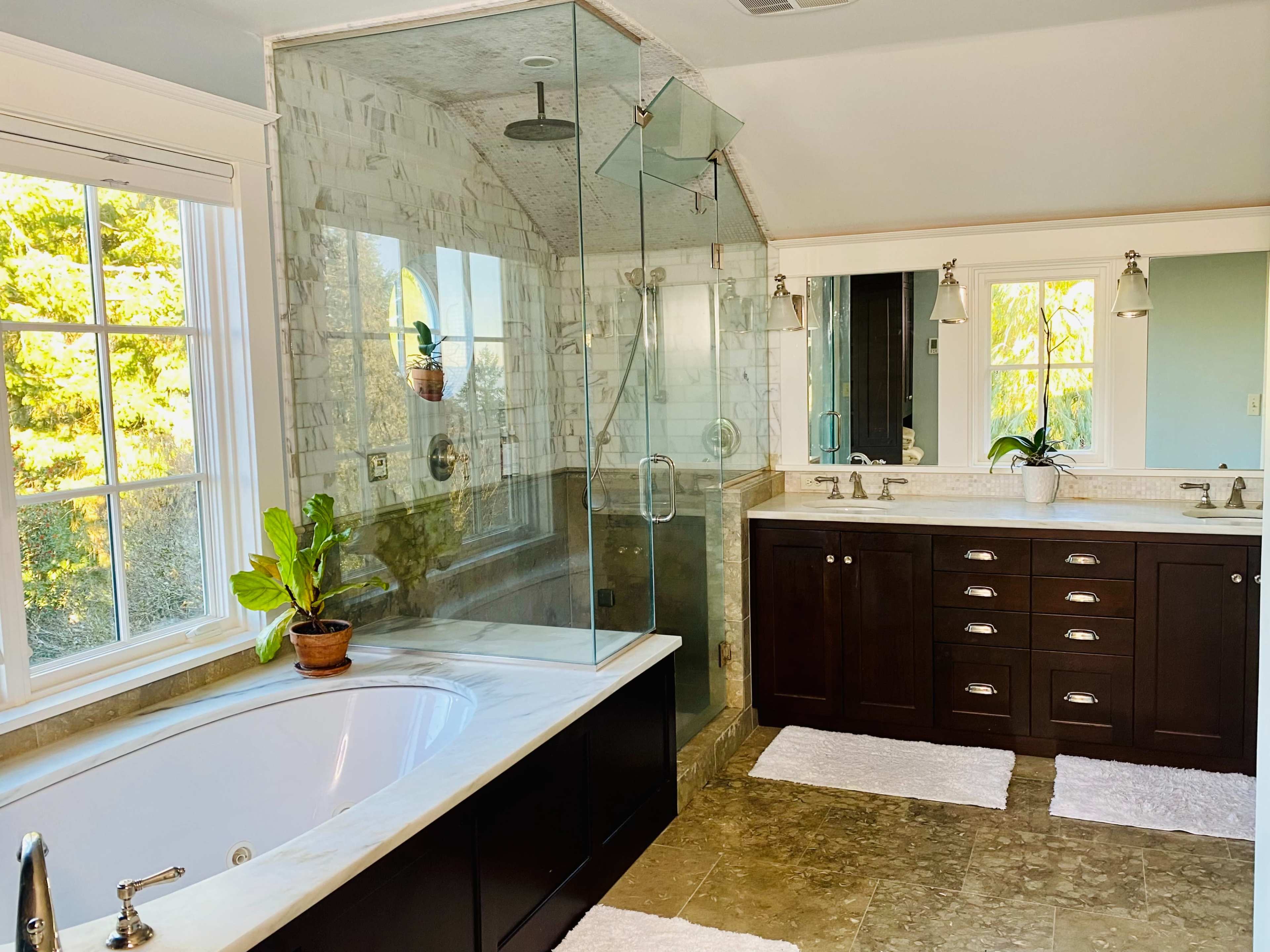 A spacious bathroom featuring a glass-enclosed shower, a freestanding bathtub, dual vanities with dark cabinetry, and large windows providing natural light.