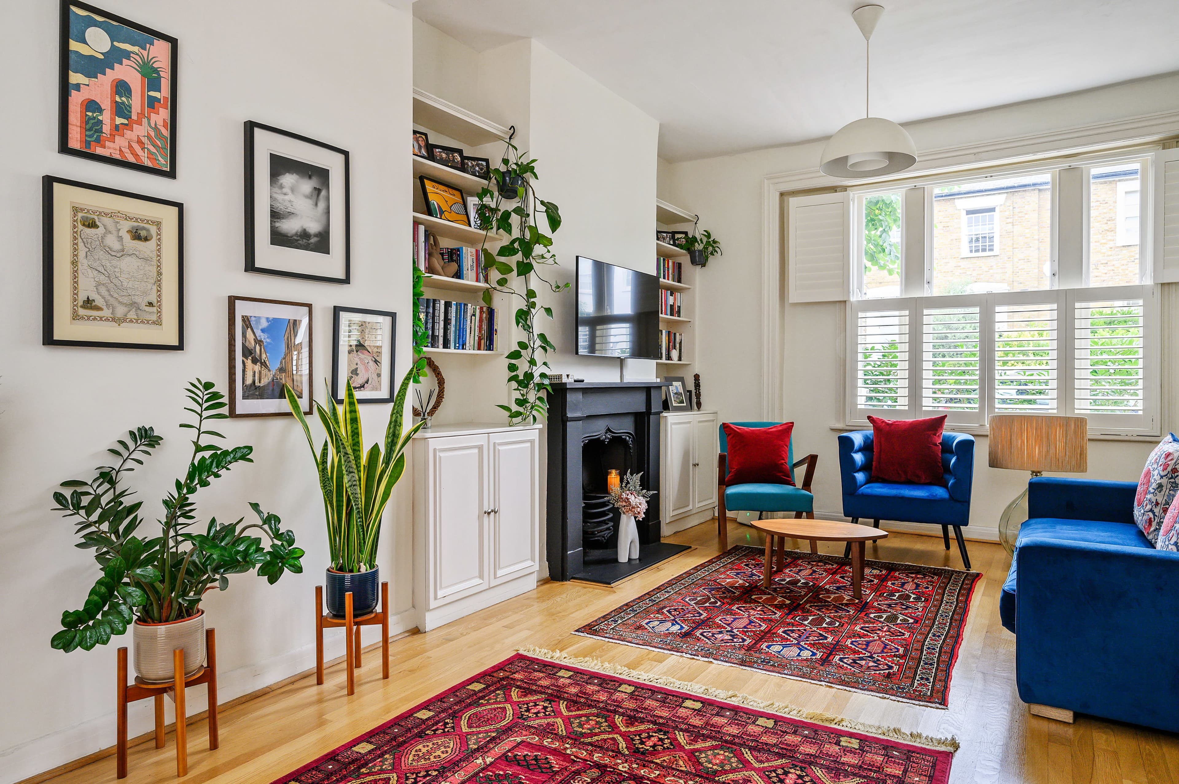 A living room features a blue sofa, two armchairs, a coffee table, shelves with books and decor, and plants, all arranged around a fireplace with a patterned rug on the floor.