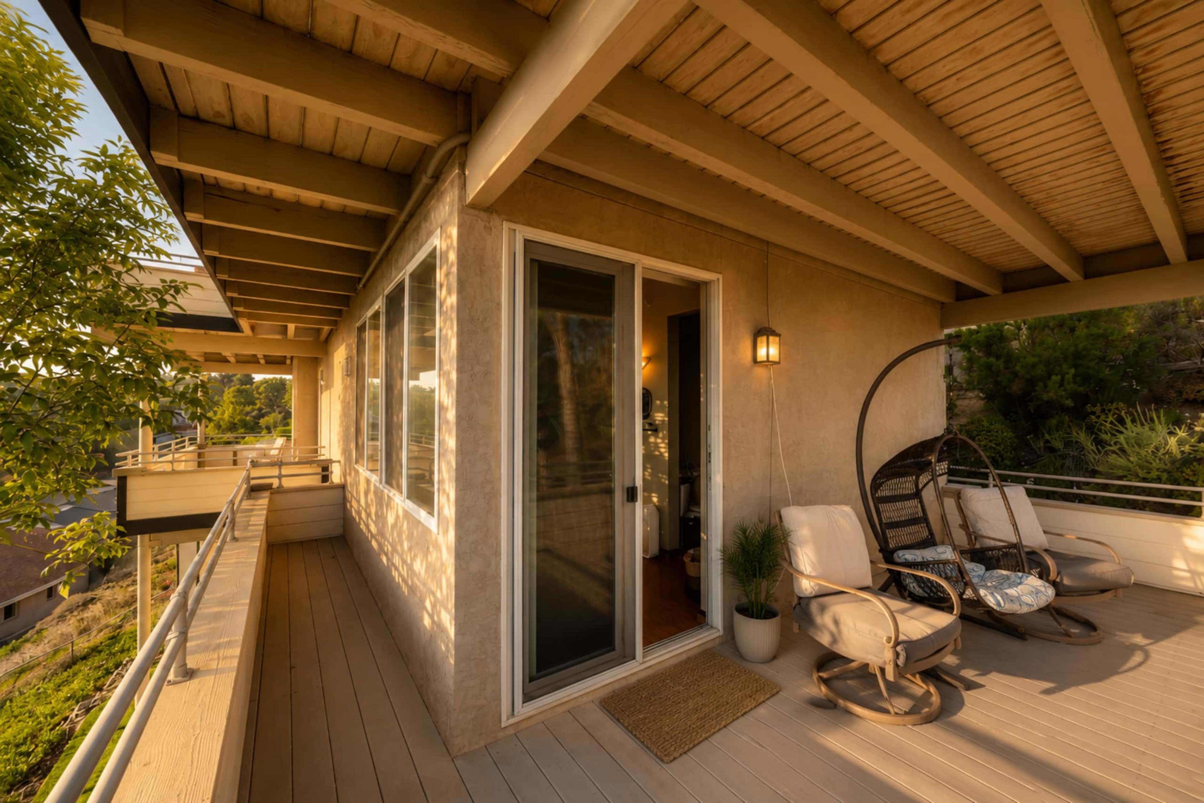 The image shows a modern balcony with two chairs and a hanging chair, featuring large windows and a door that opens into an interior space.