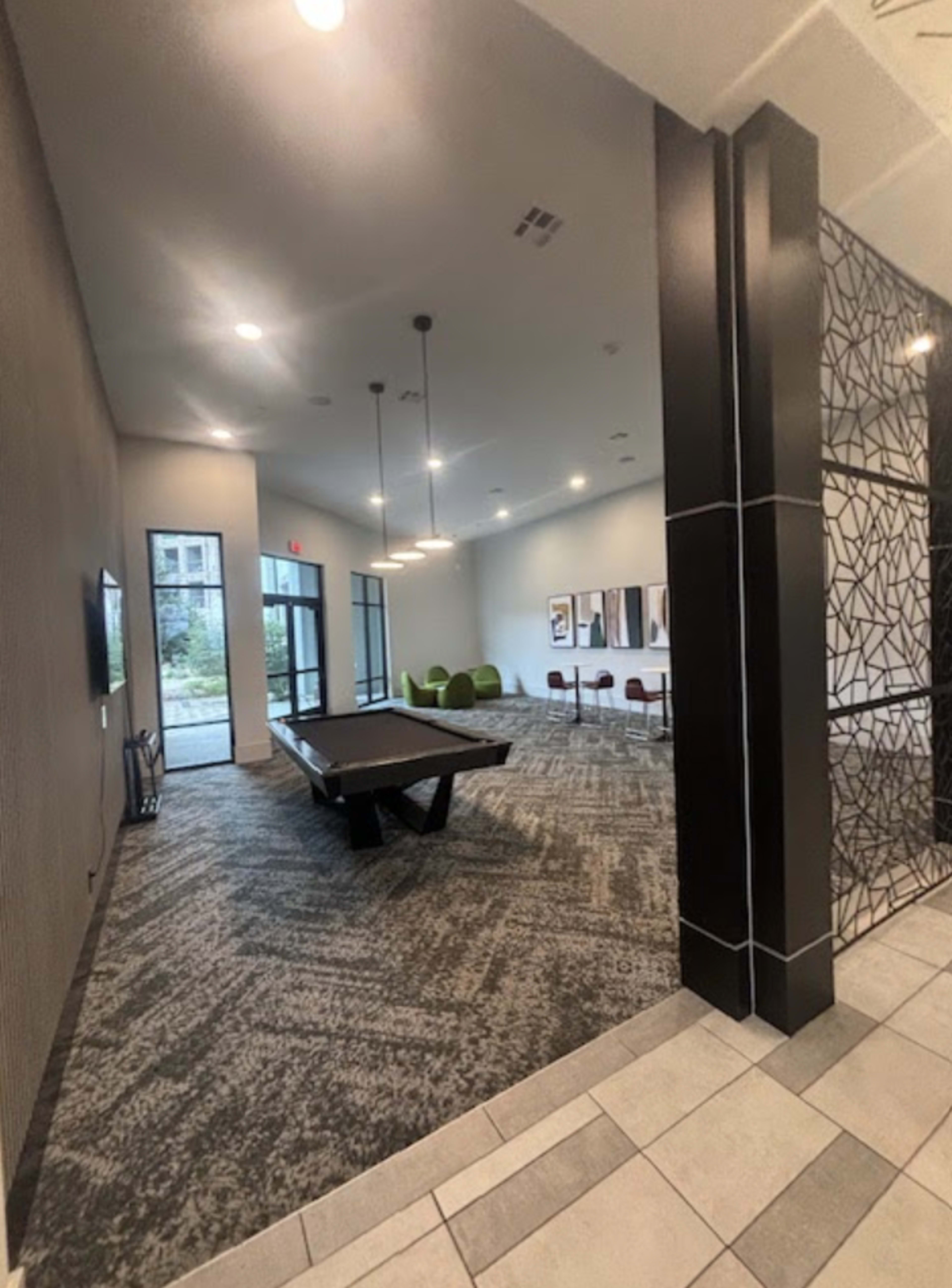 The image shows a modern communal lounge area featuring a pool table, seating, and an entrance to the outdoors.