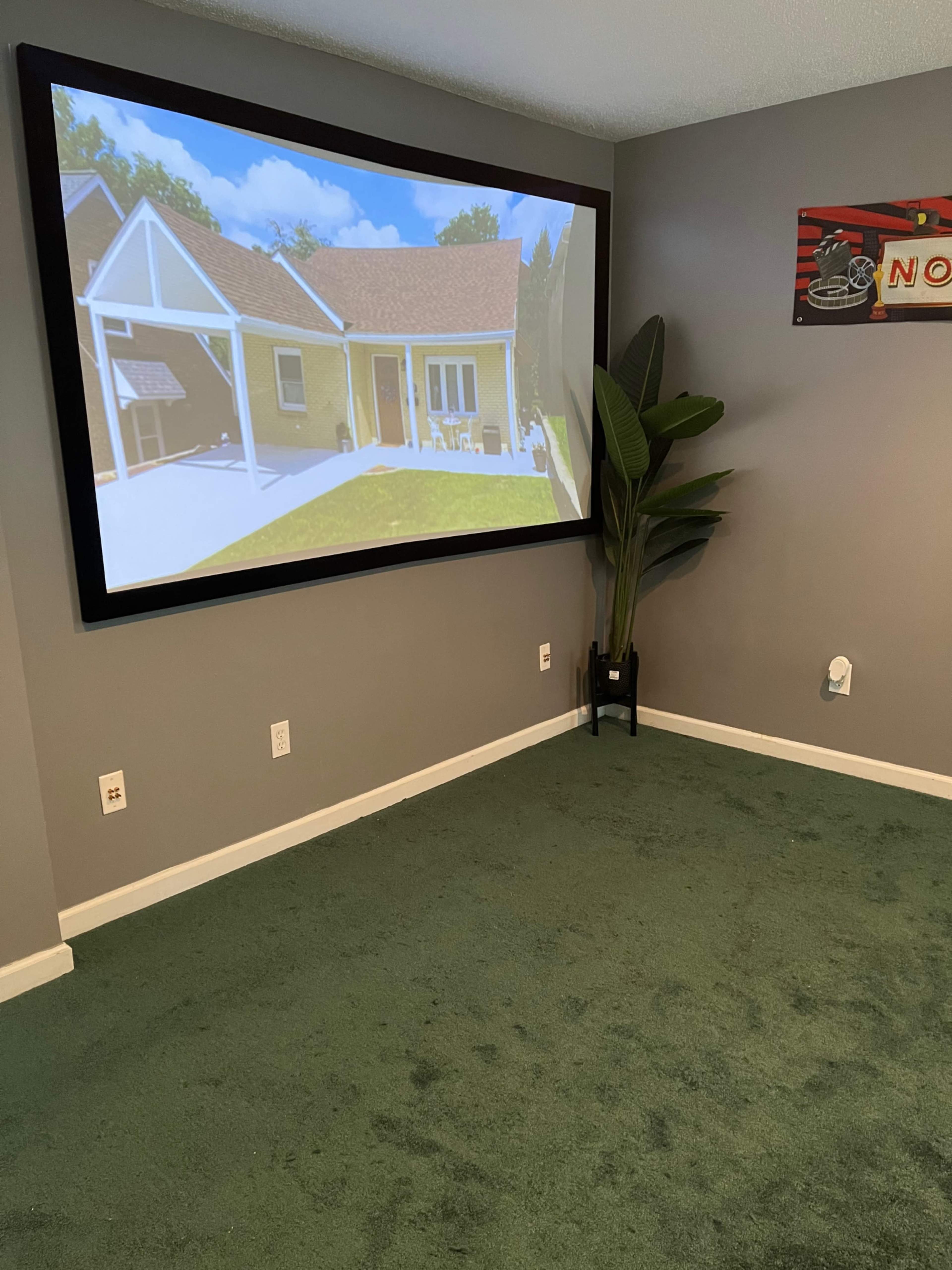 A large projector screen displaying an image of a house is mounted on a gray wall, with a green carpet and a potted plant nearby.