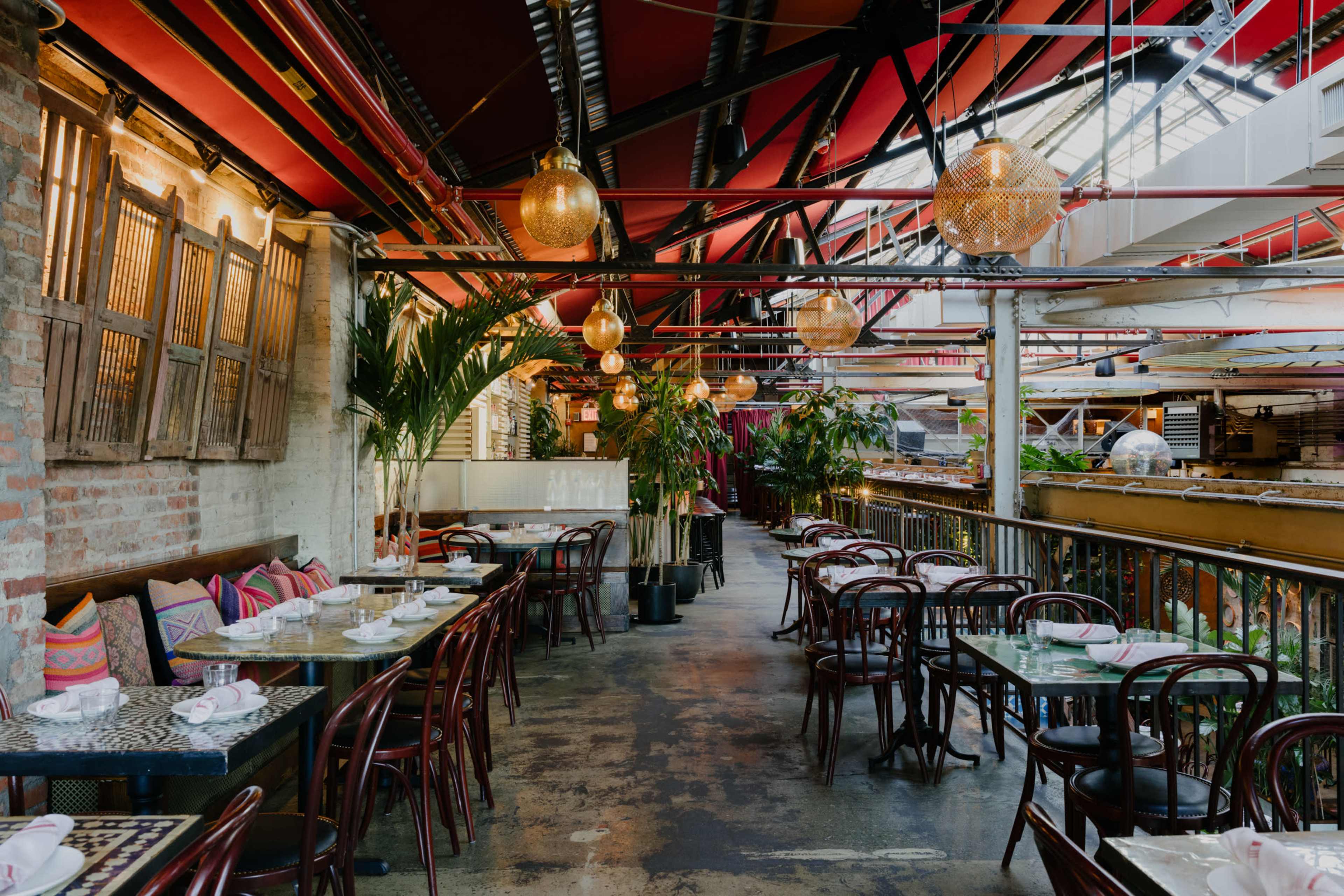 An indoor restaurant with wooden tables, chairs, and green plants, set under red beams and hanging light fixtures.