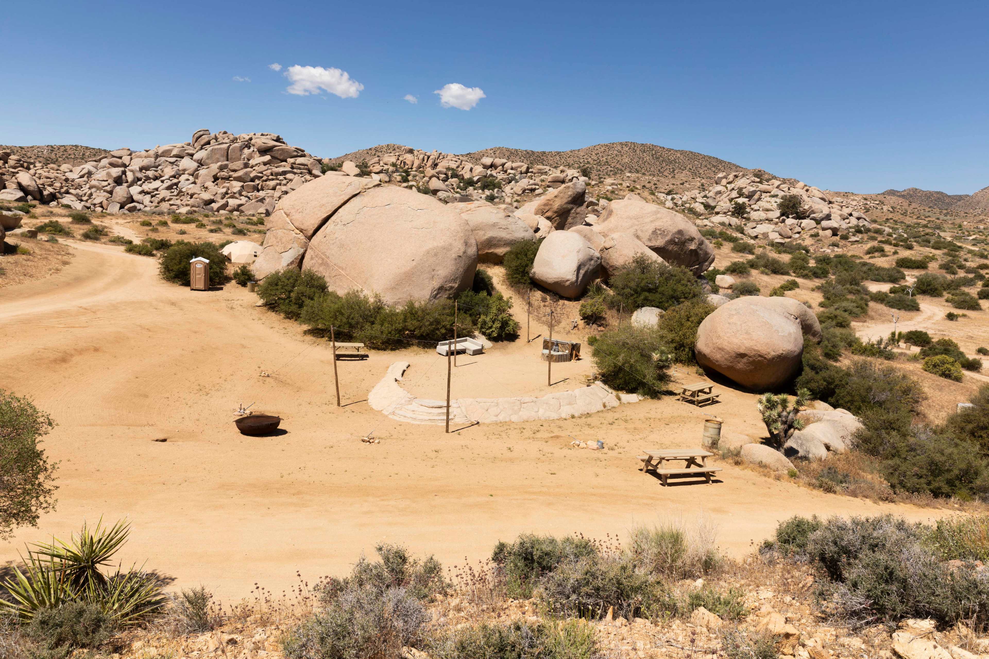 A desert landscape with large boulders, a dirt path, and several signs near a seating area surrounded by sparse vegetation.