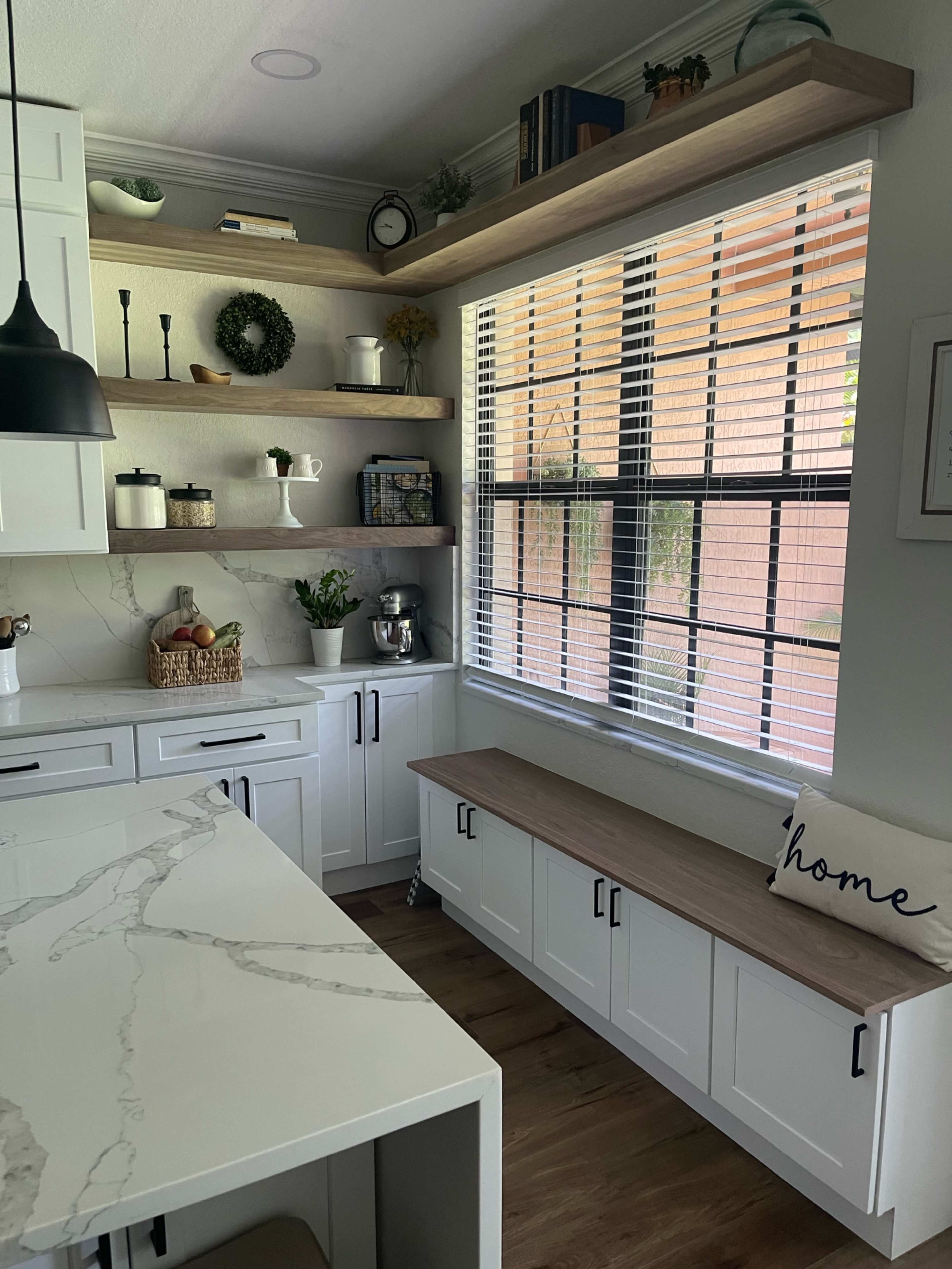 A modern kitchen features white cabinets, a marble countertop, open shelving with decorative items, and a wooden bench by a window.