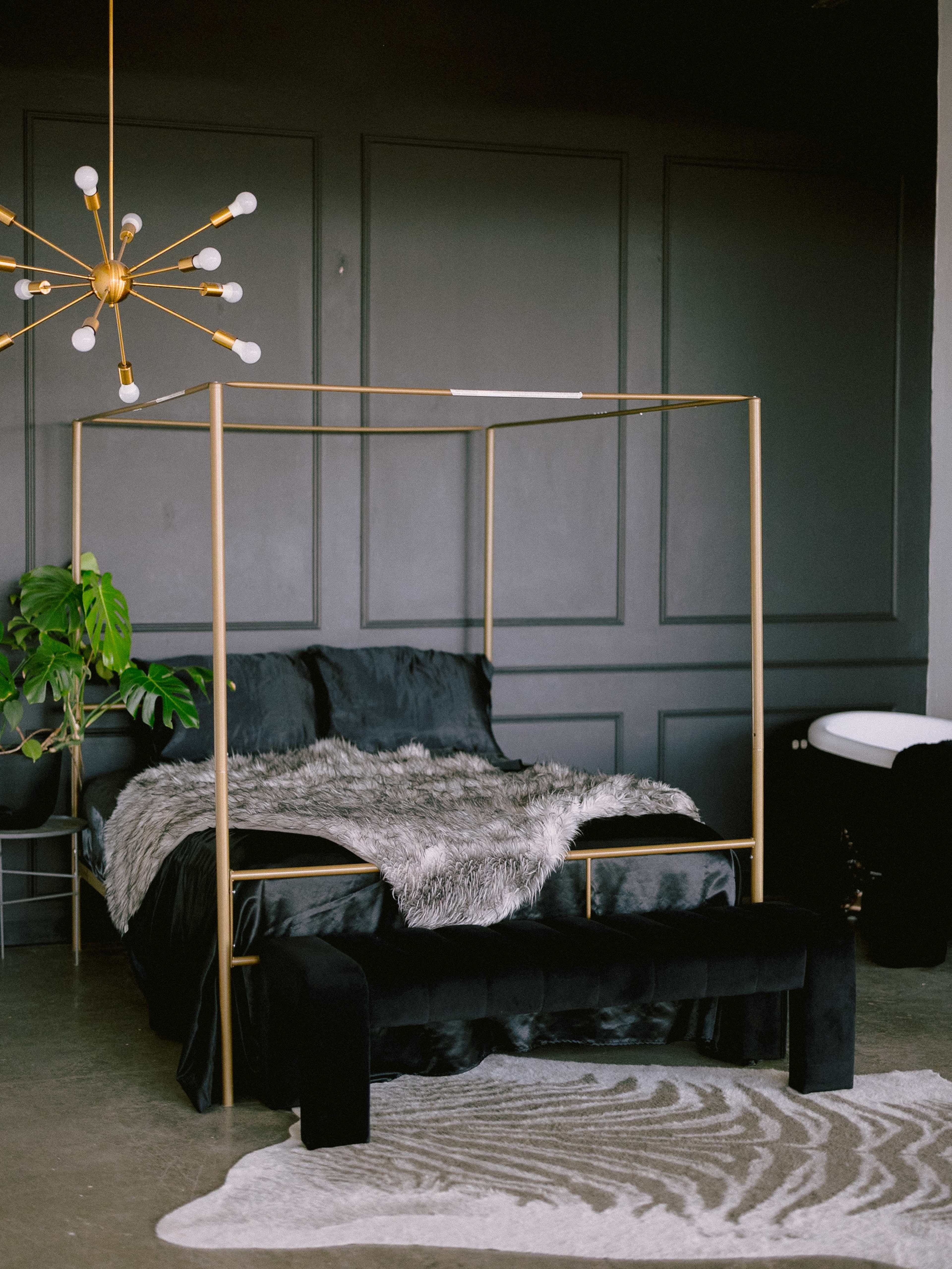 The image shows a modern bedroom with a gold-framed canopy bed, black bedding, a large faux fur throw, a geometric light fixture, and a patterned area rug on the floor.