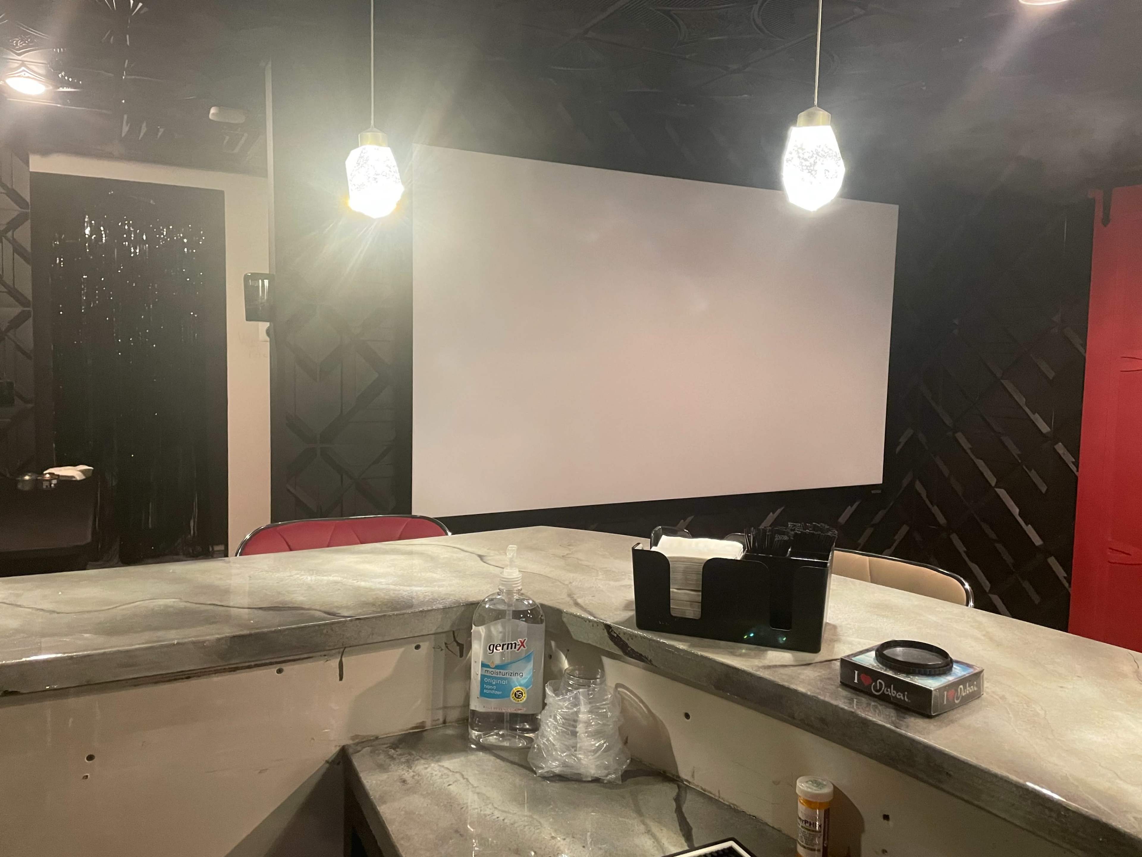 A dimly lit room with a large blank screen on one wall, a bar counter with various items, and pendant lights above.