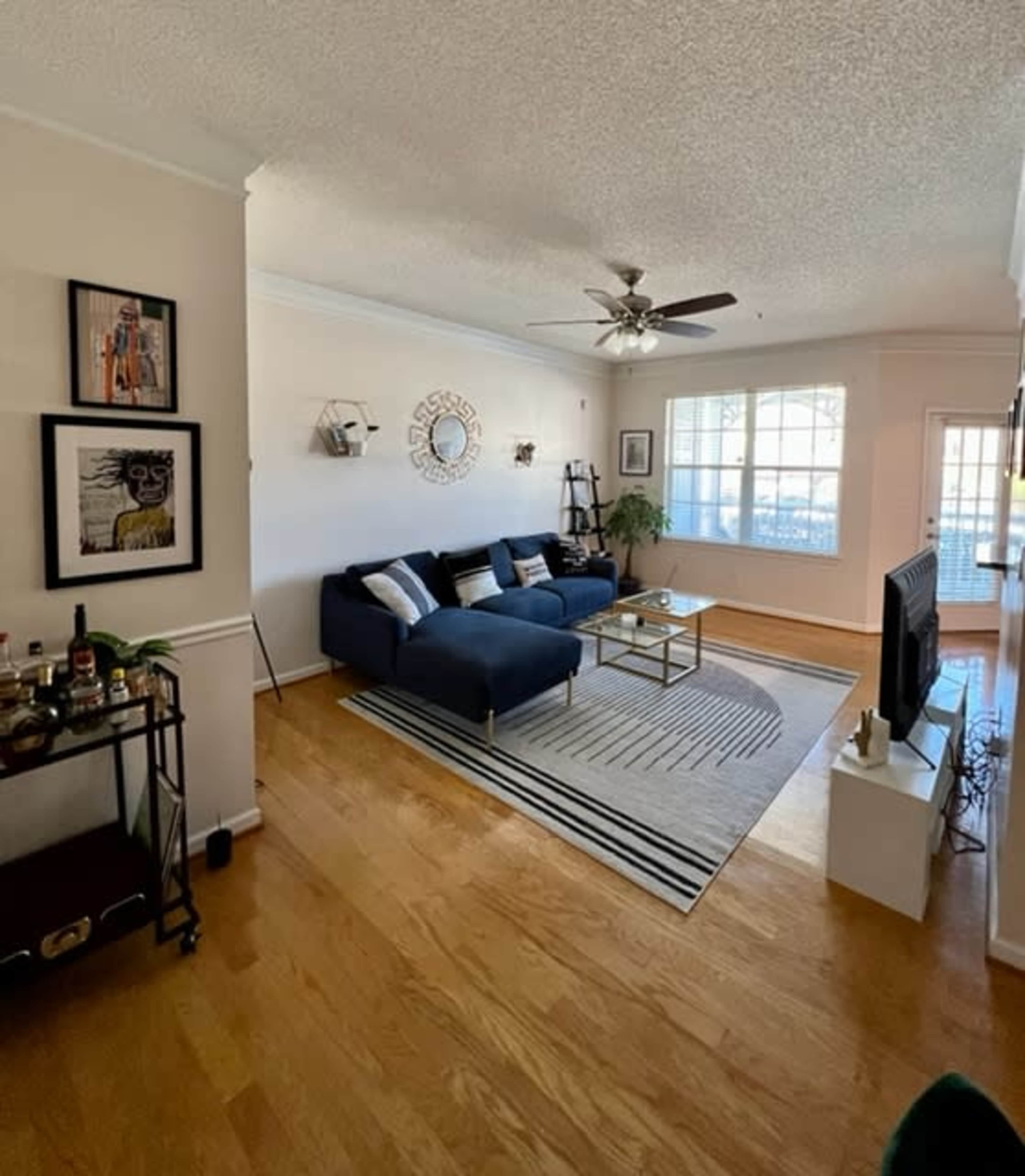 A living room featuring a blue sofa, a glass coffee table, and a wooden floor, with a bar cart in one corner and large windows providing natural light.