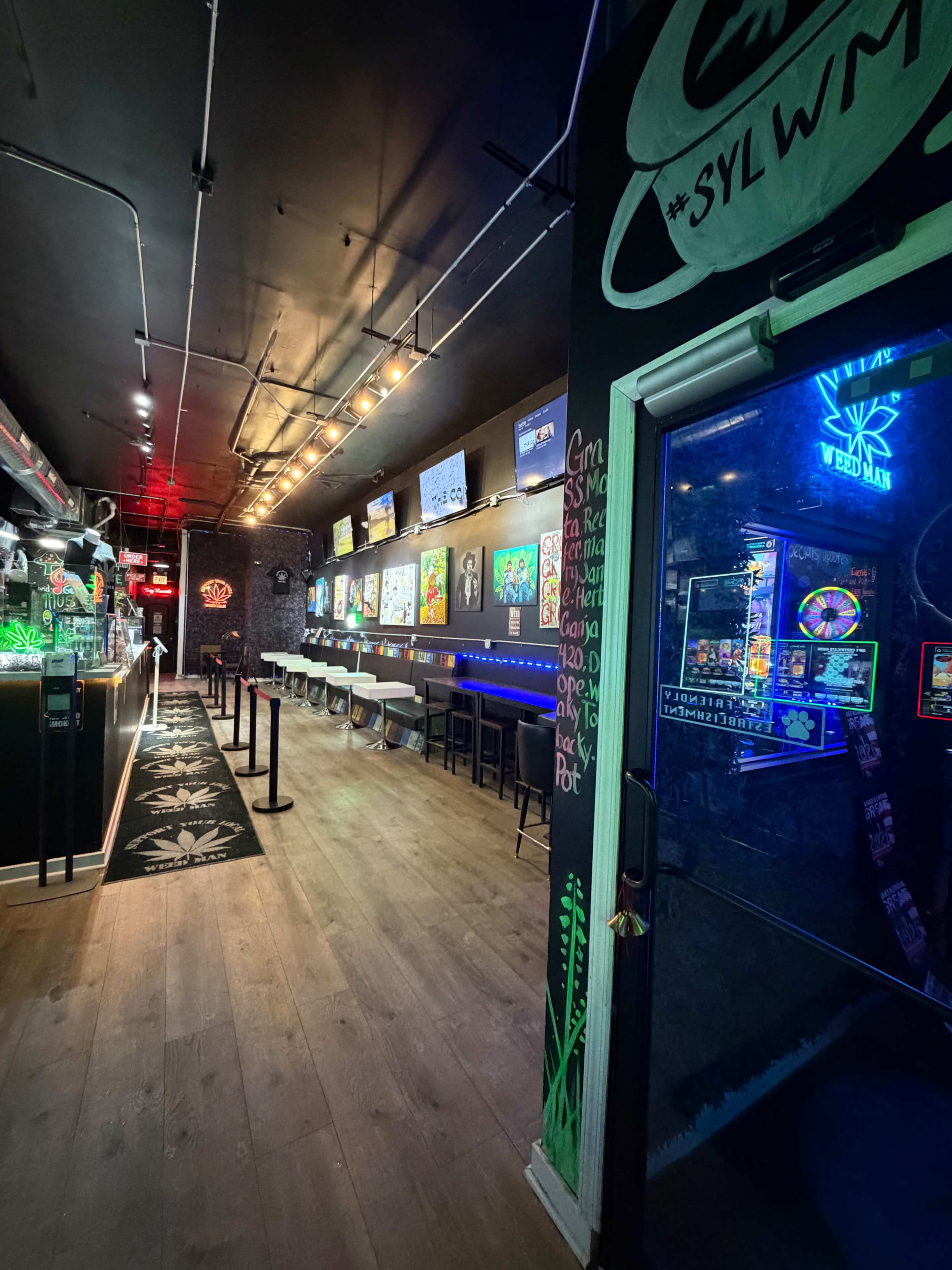 The image shows a dimly lit bar with colorful artwork on the walls, a long counter with seating, and a pathway leading towards the entrance.