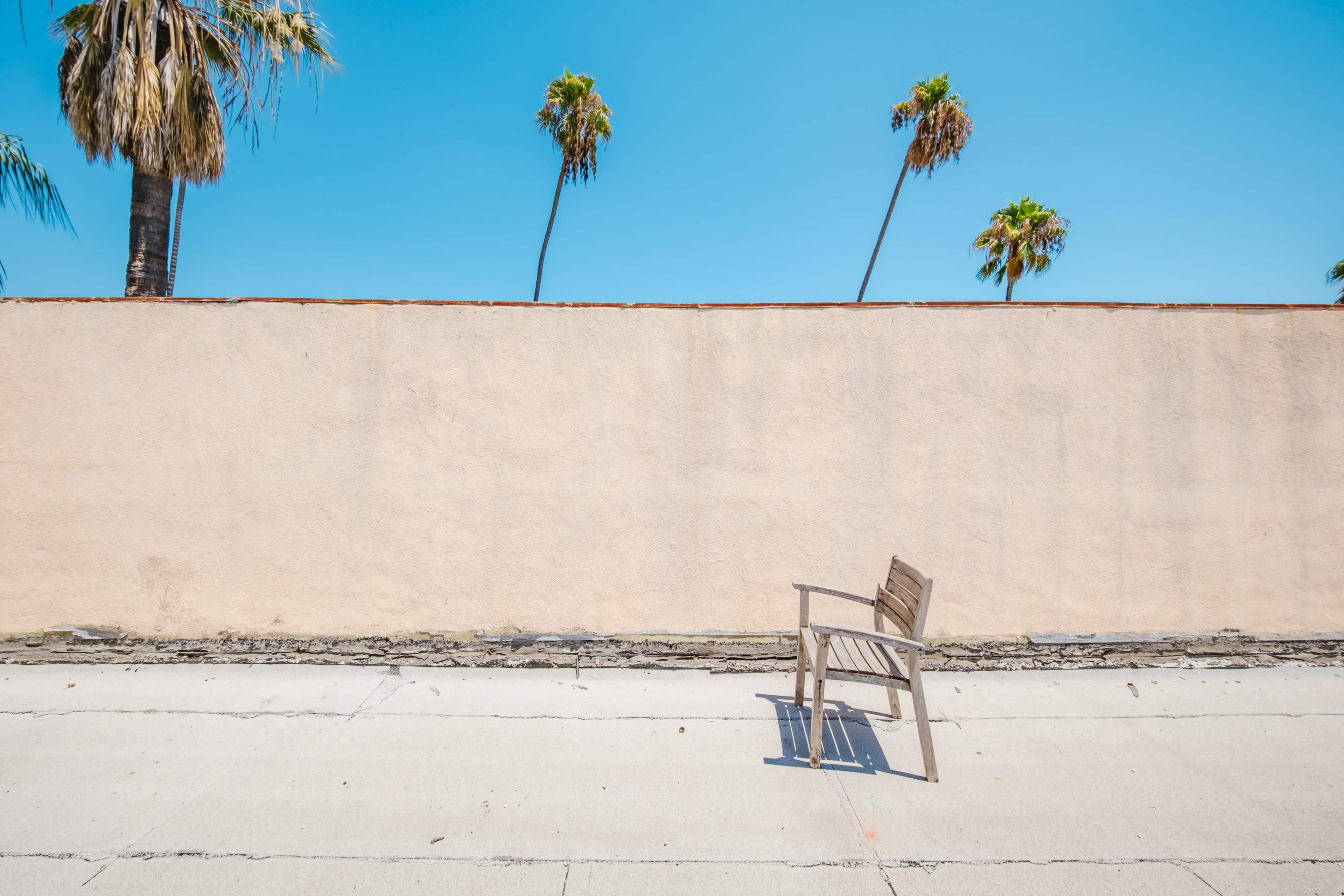 A wooden chair is positioned on a concrete surface in front of a bare wall, with palm trees visible against a clear blue sky.