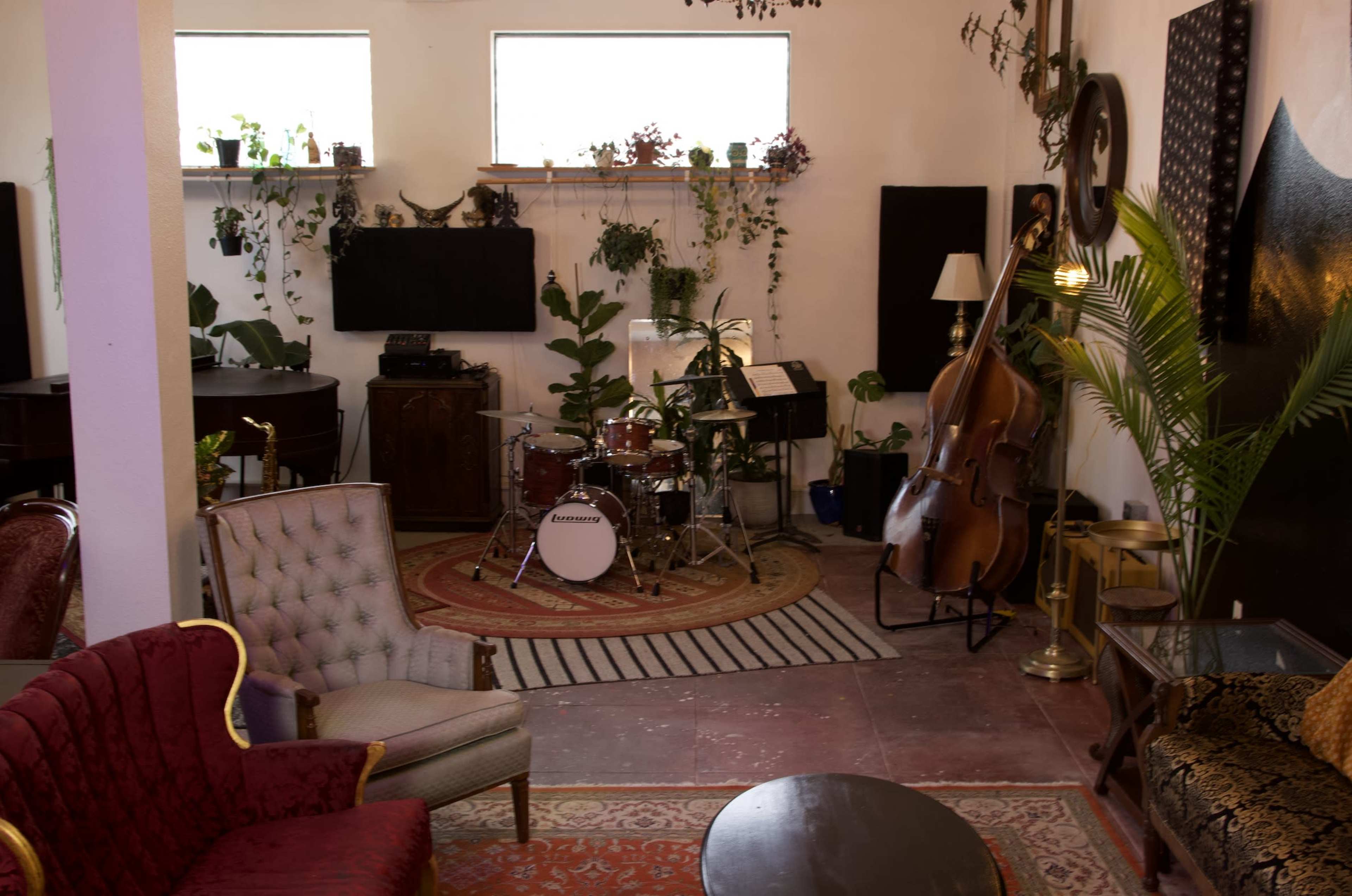 The interior of a music studio features a drum set, a double bass, potted plants, and various seating areas.
