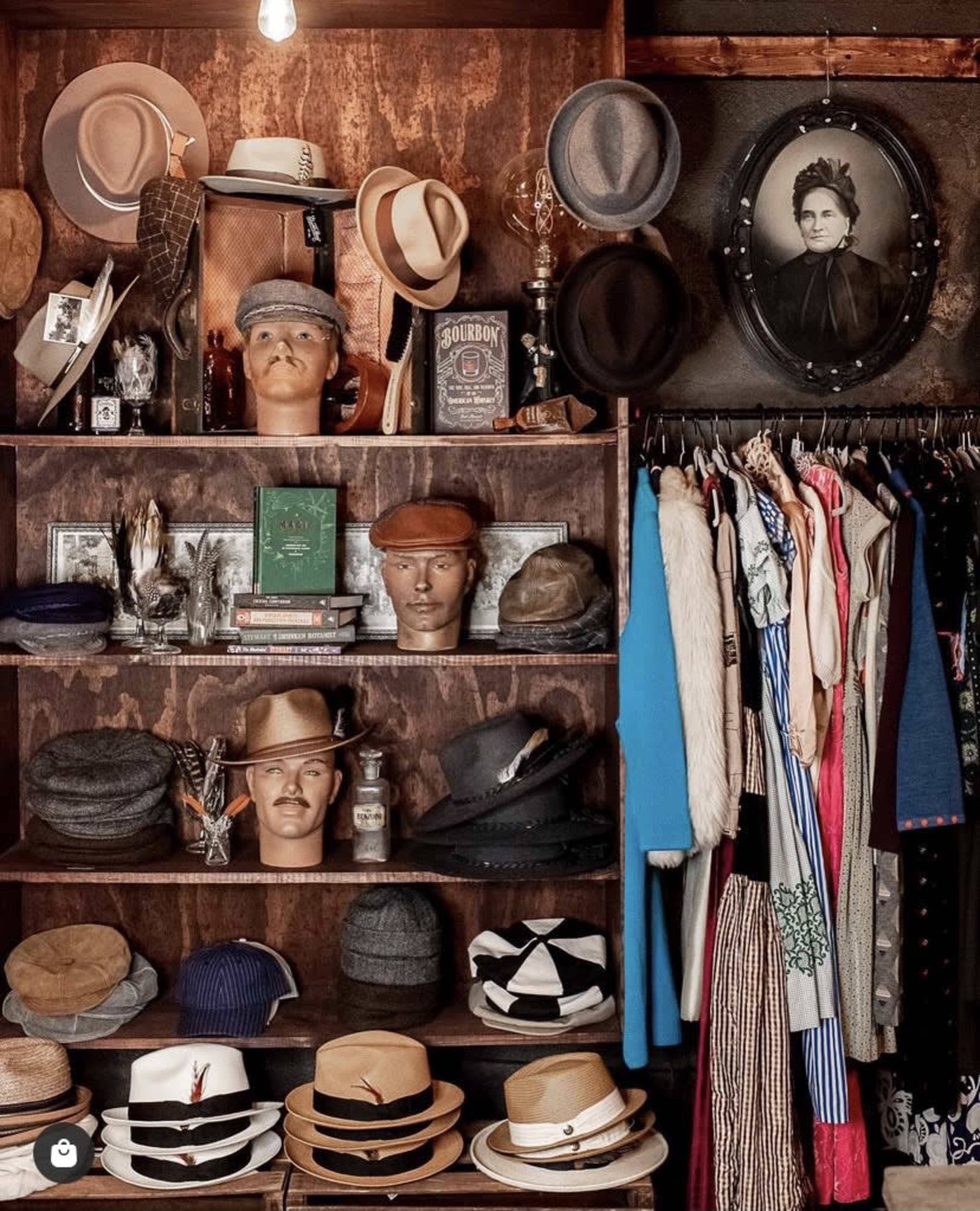 A vintage display featuring various hats arranged on mannequins and shelves, alongside clothing items hanging on a rack and a framed portrait on the wall.