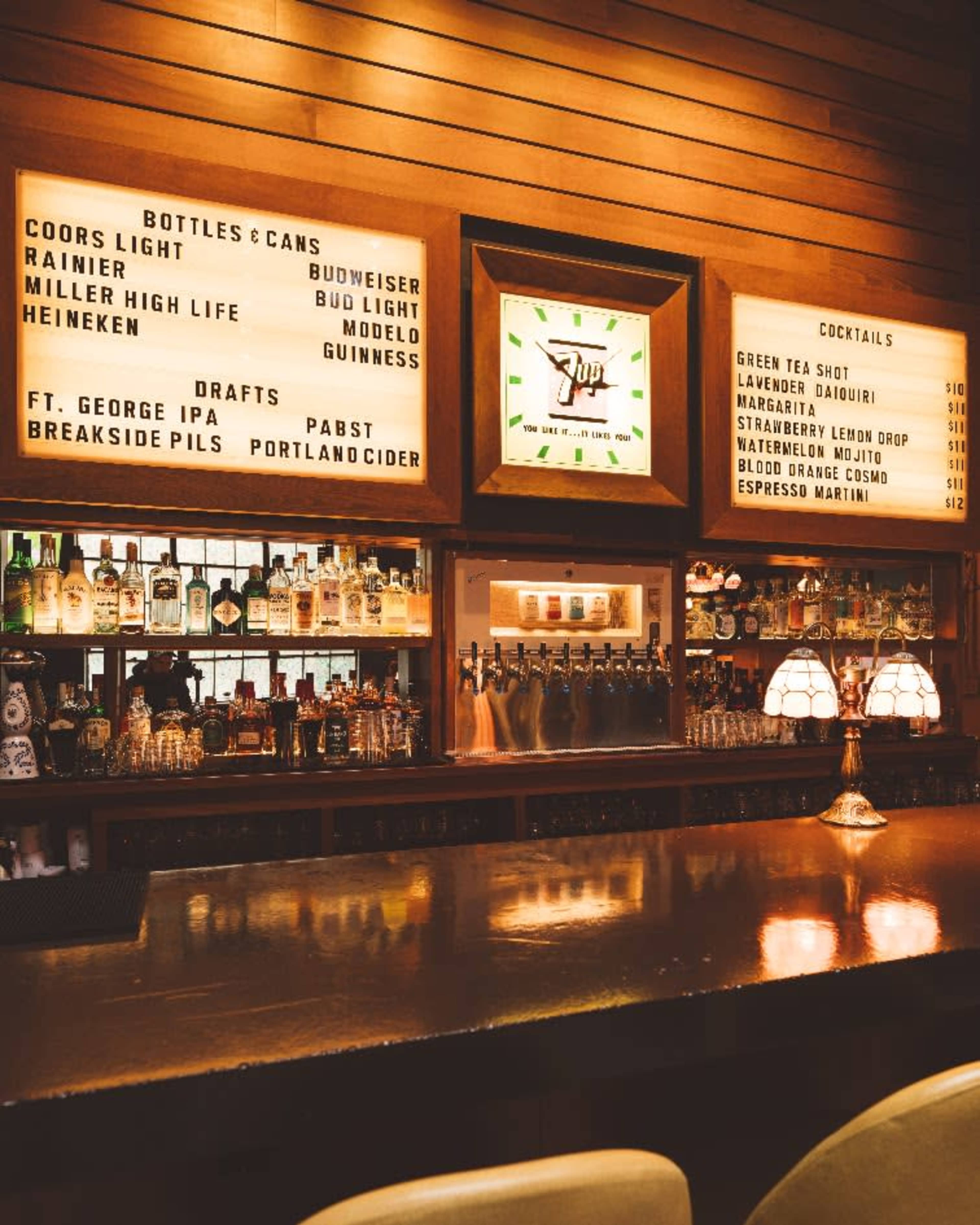 The image shows a well-lit bar with a wooden counter, shelves lined with various liquor bottles, and a menu listing beer and cocktail options displayed on a backboard.