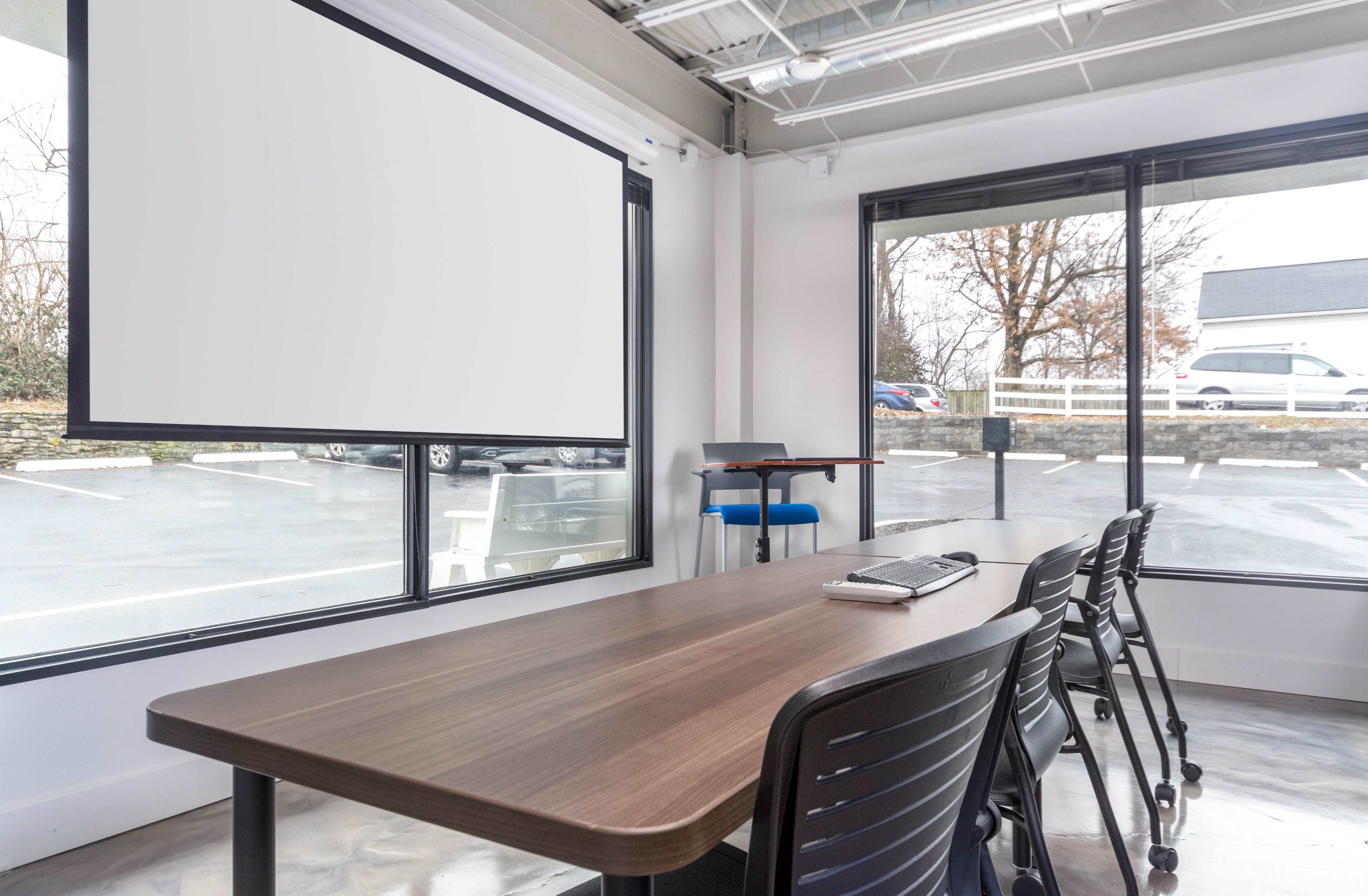 A conference room features a large table, black chairs, a projector screen, and large windows overlooking a parking lot.