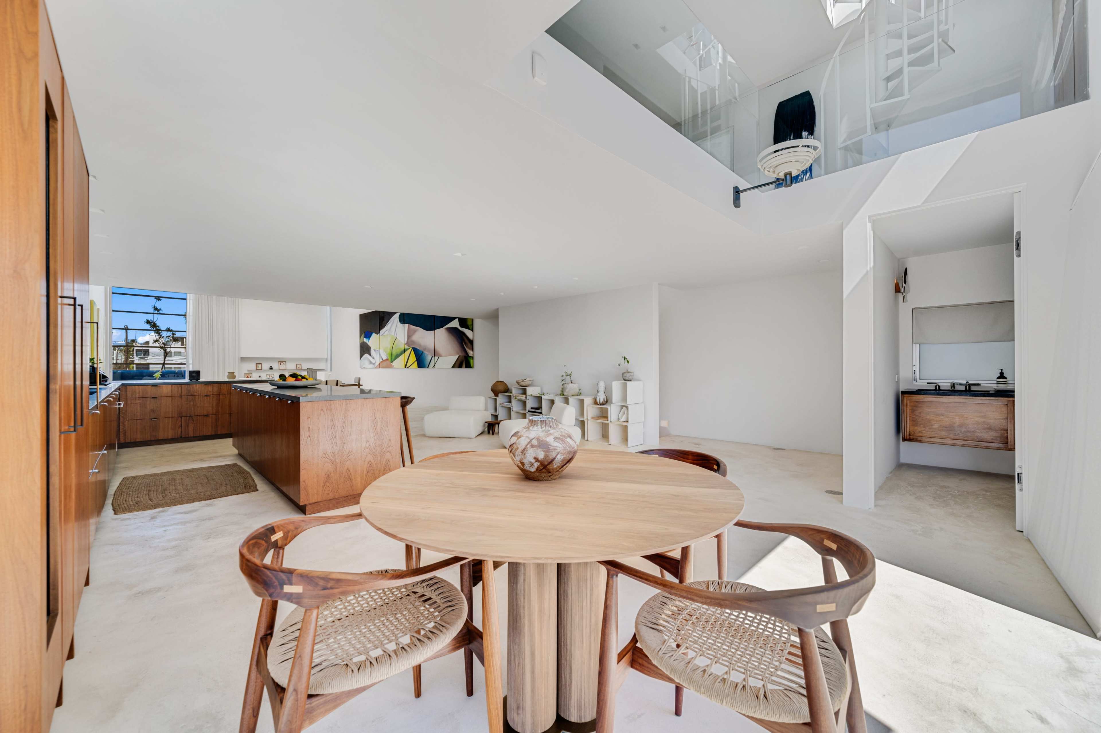 The image shows a modern, open-concept kitchen and dining area featuring a round wooden table with woven chairs, a sleek kitchen with cabinetry, and a light-filled living space.