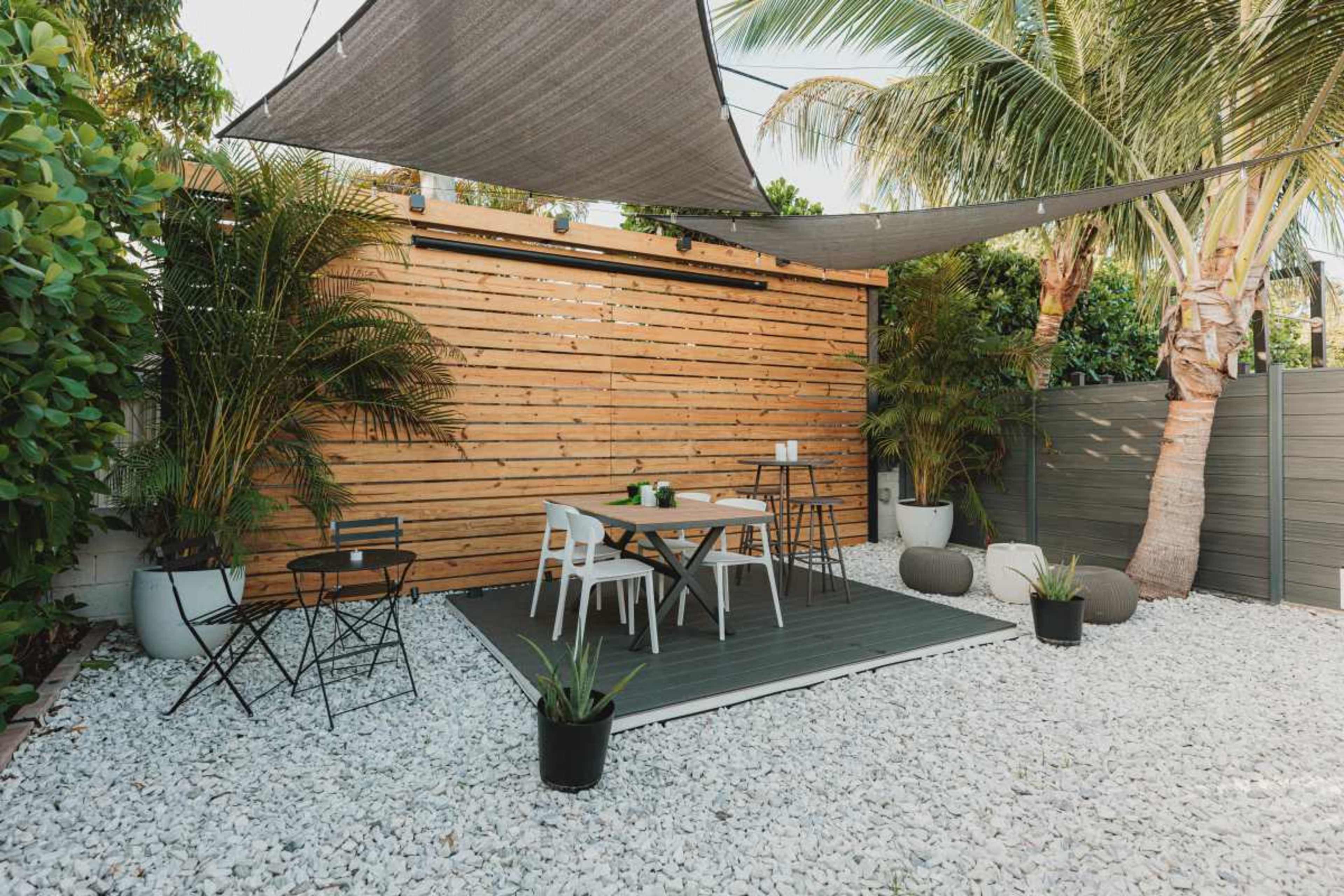 The image shows a patio area with a wooden slatted wall, a table and chairs, and surrounding greenery, all set on a bed of white gravel.
