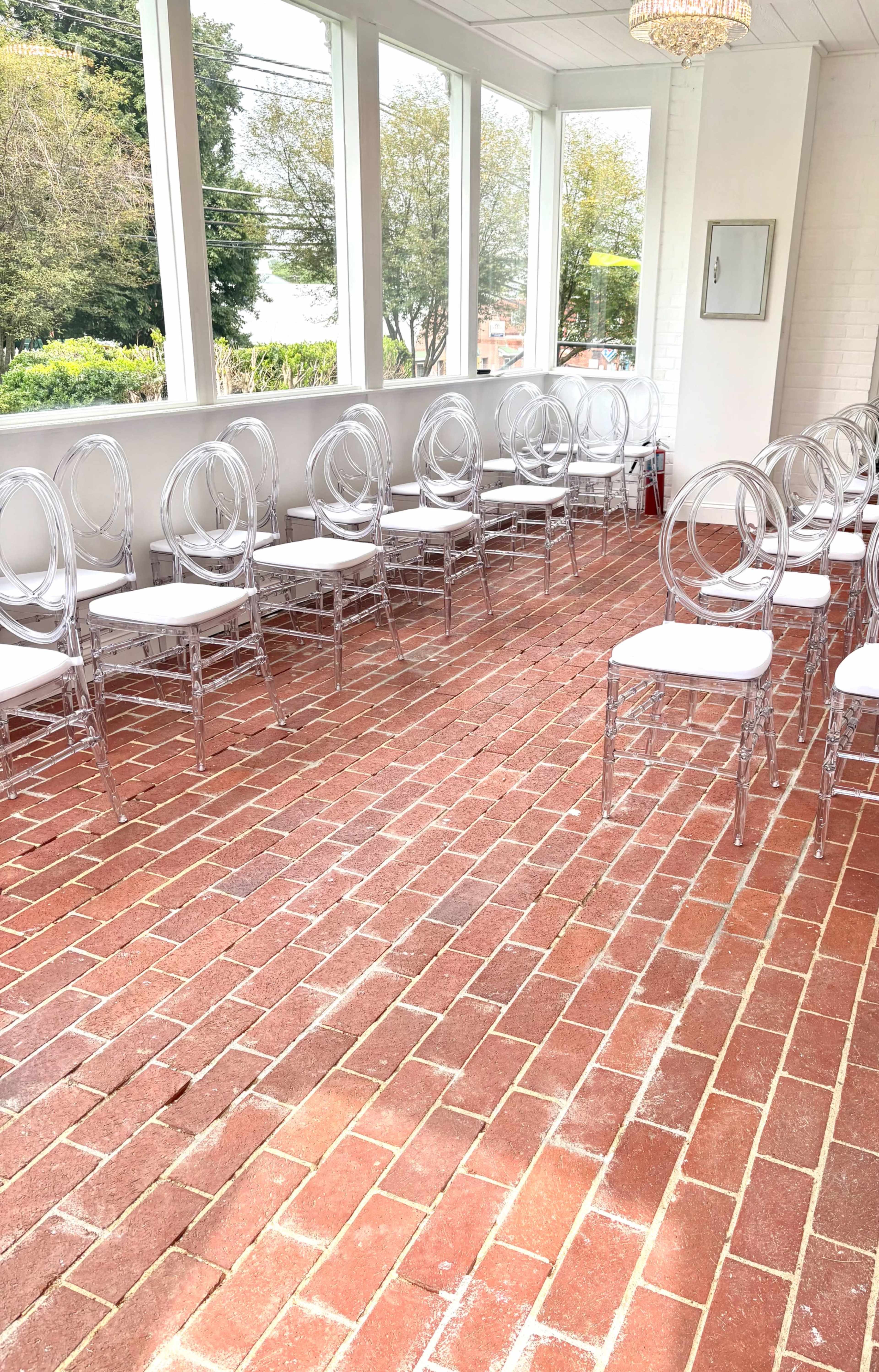 The image shows a sunlit indoor space with a brick floor and several clear acrylic chairs arranged in two rows.