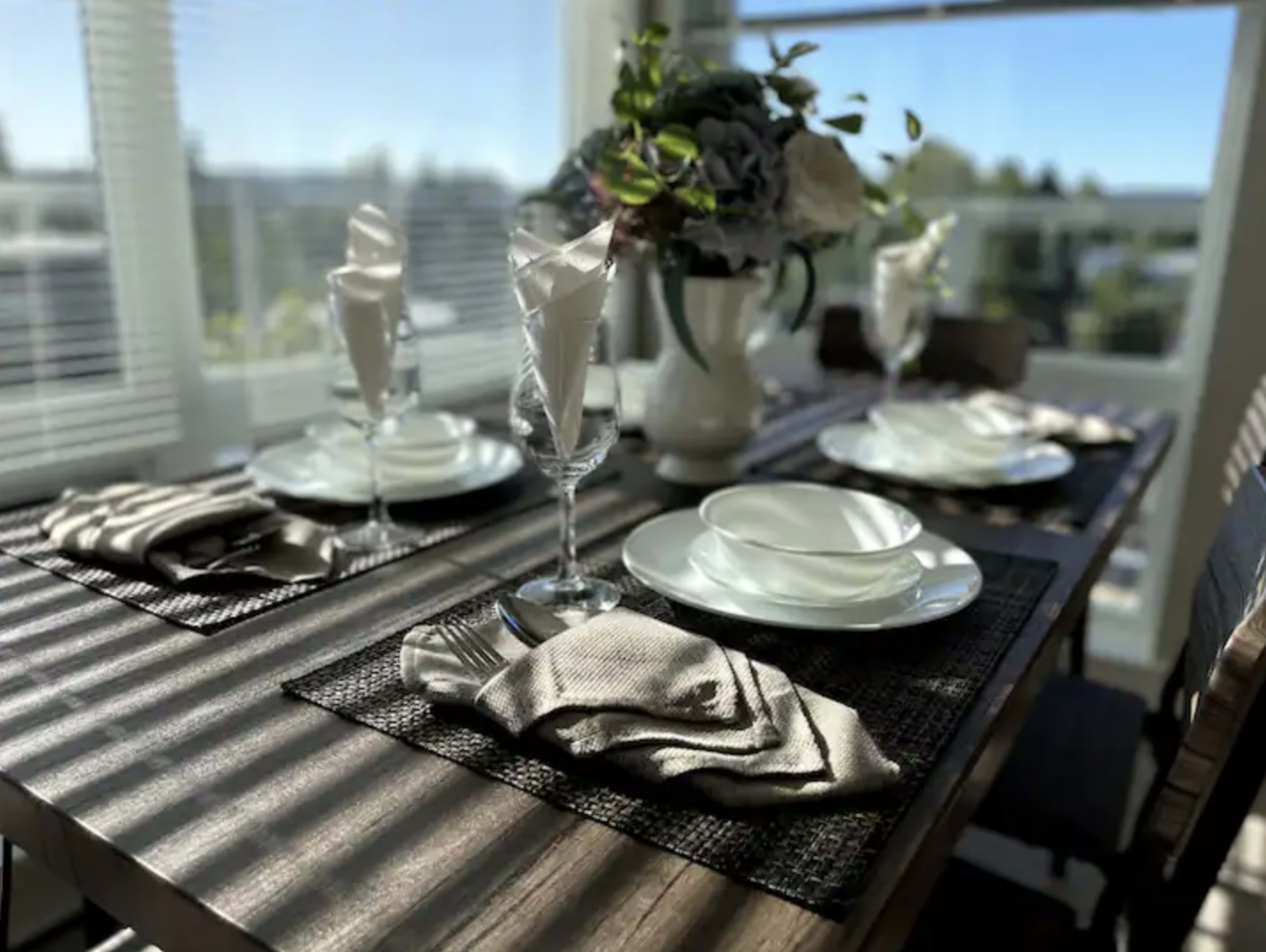 A dining table is set with glassware, plates, and neatly folded napkins, surrounded by a floral centerpiece and illuminated by natural light.