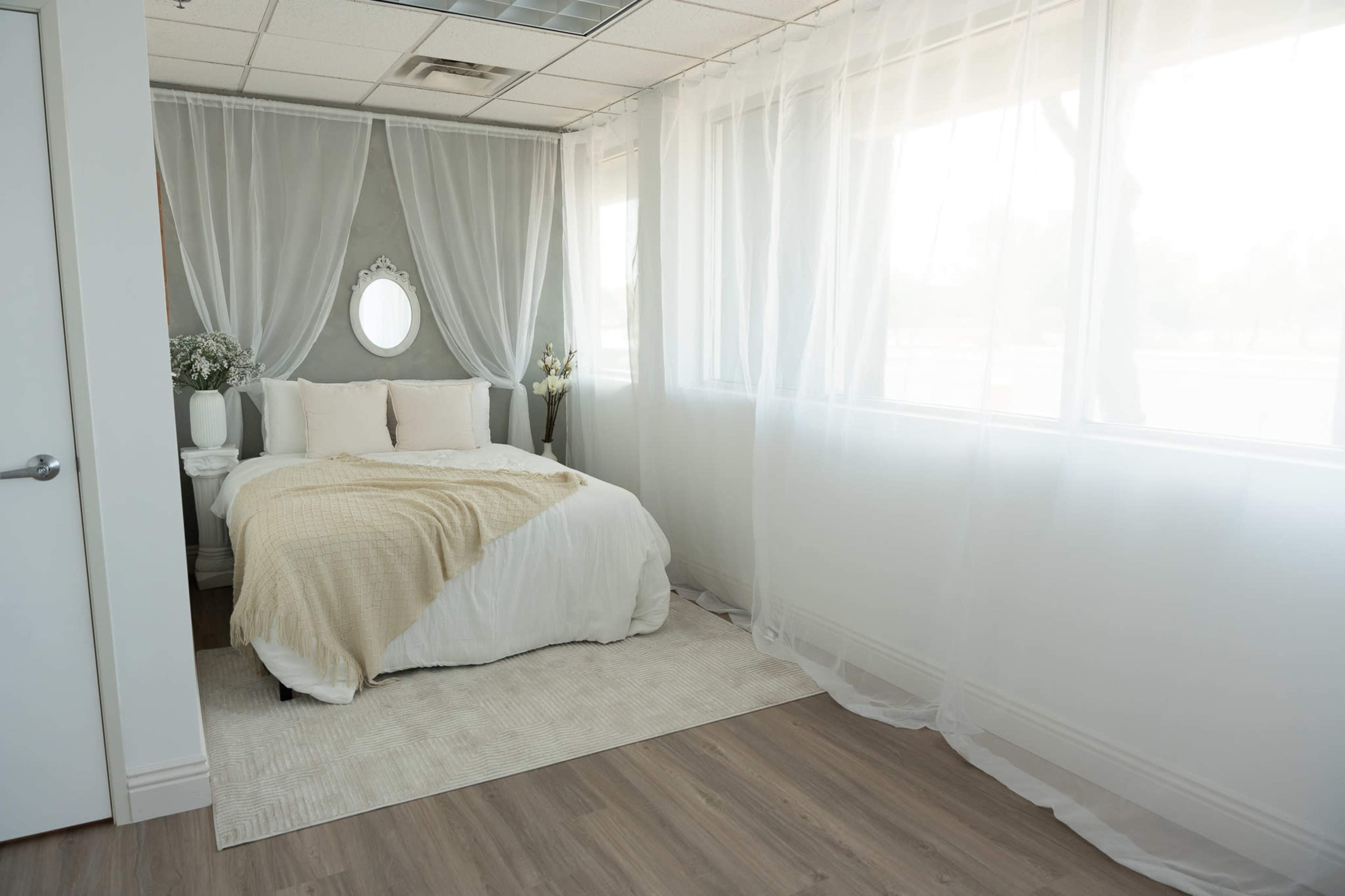 A softly lit bedroom features a bed with white and beige bedding, surrounded by sheer curtains and a decorative mirror.