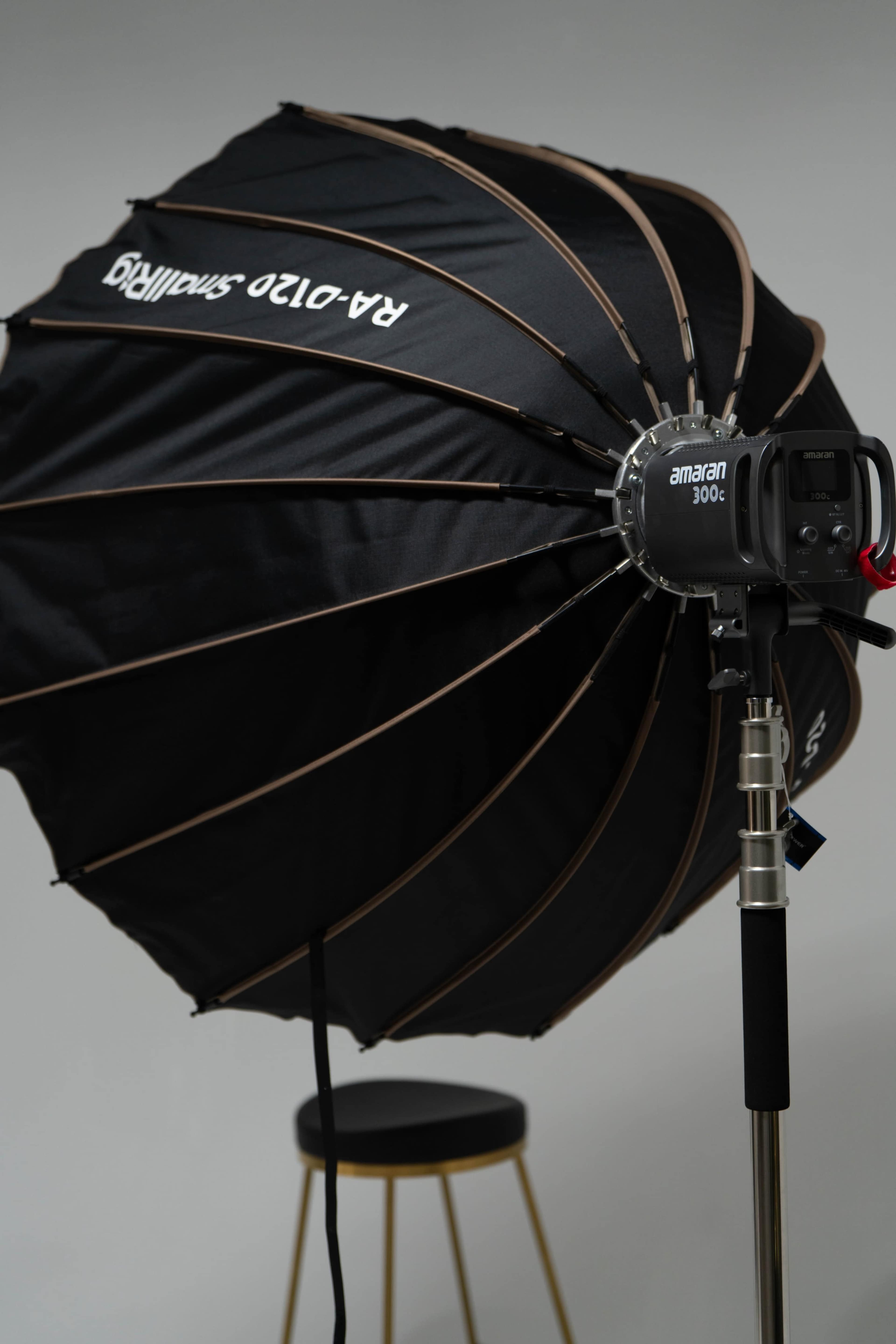 A black umbrella softbox with a strobe light attached is positioned beside a gold stool against a neutral backdrop.