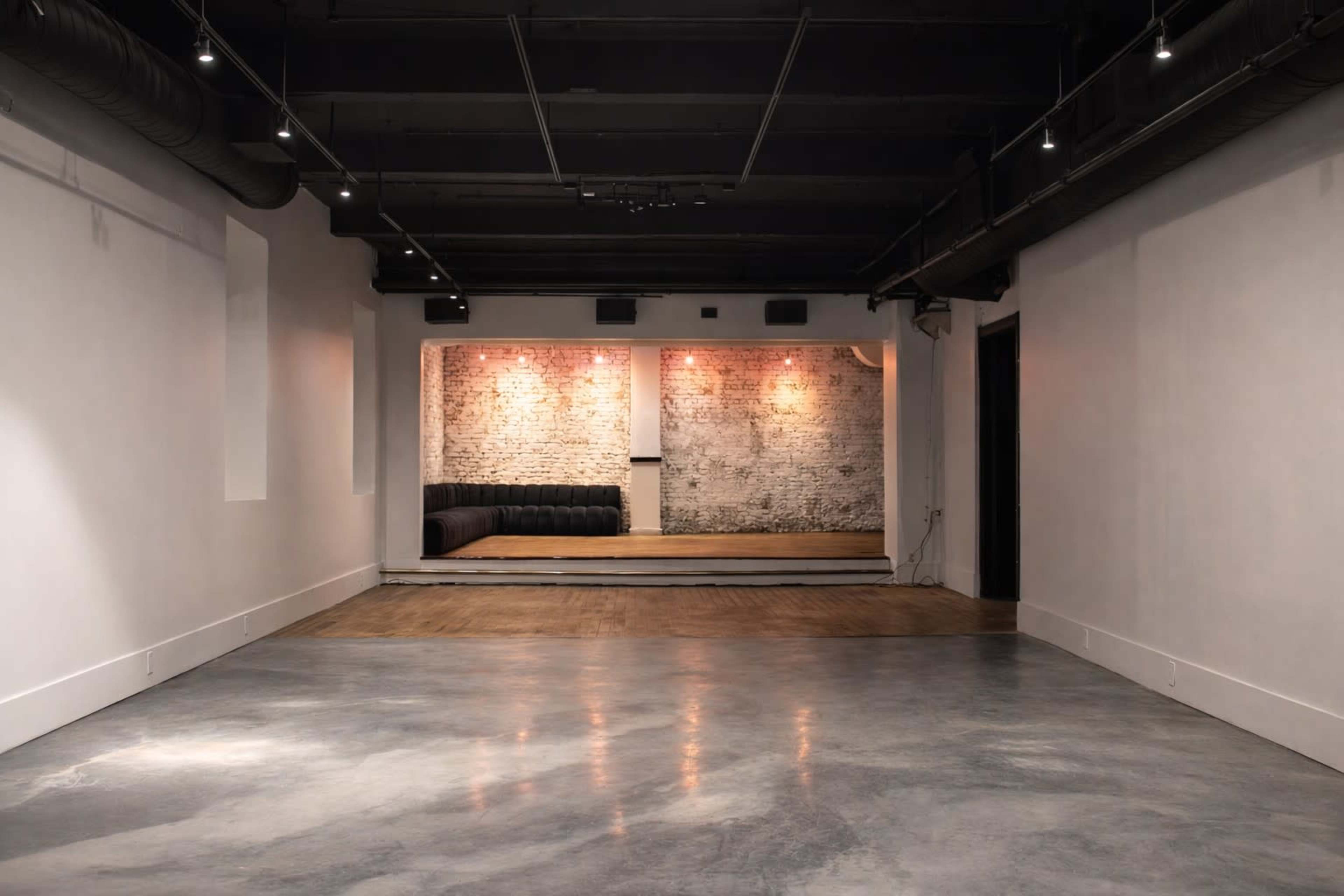 The image shows a spacious, minimally furnished room with a concrete floor and exposed brick walls, featuring a cozy seating area highlighted by soft lighting.