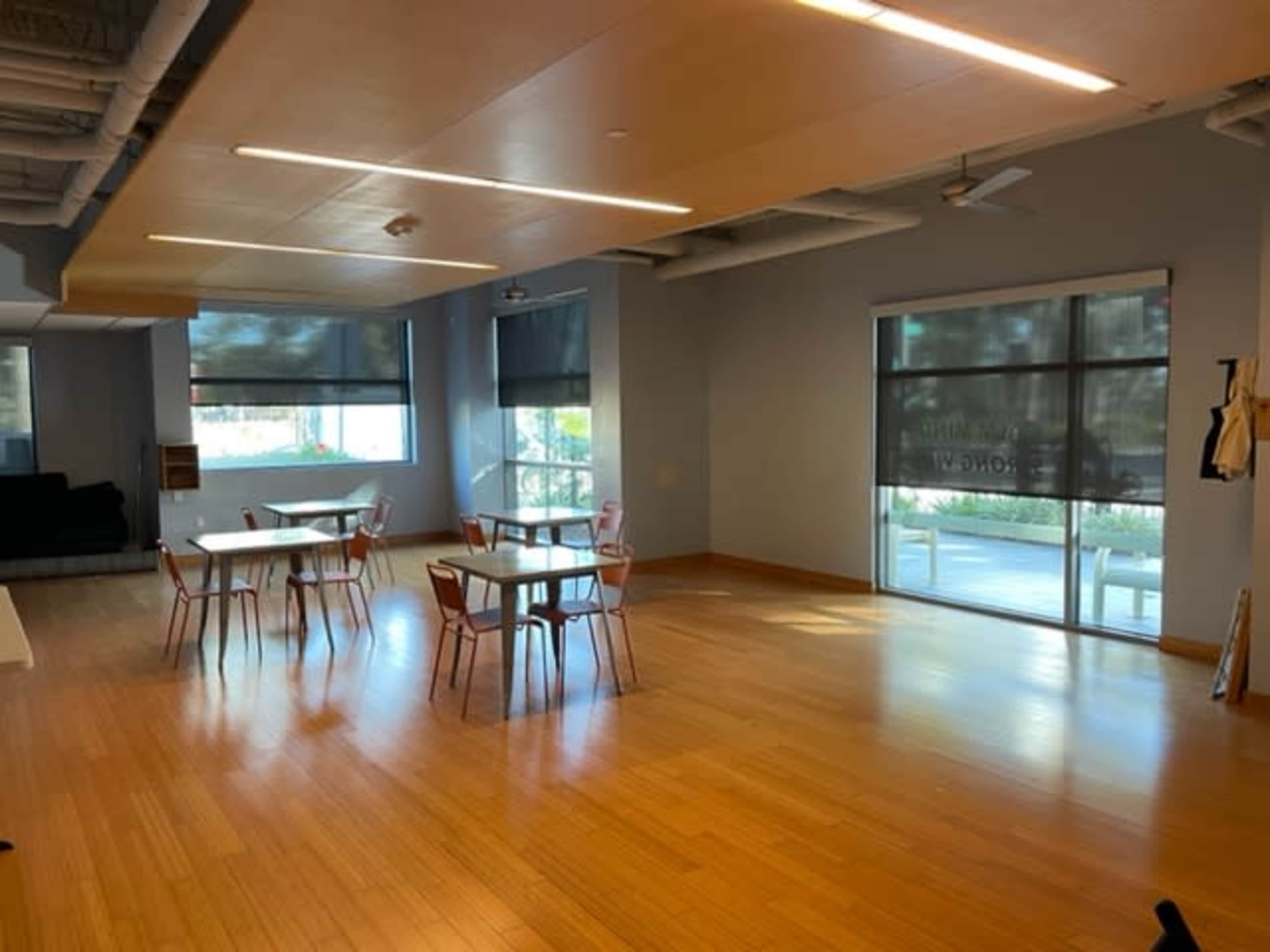 A spacious room with wooden floors, several tables surrounded by chairs, large windows with shades, and a view of the outdoors.