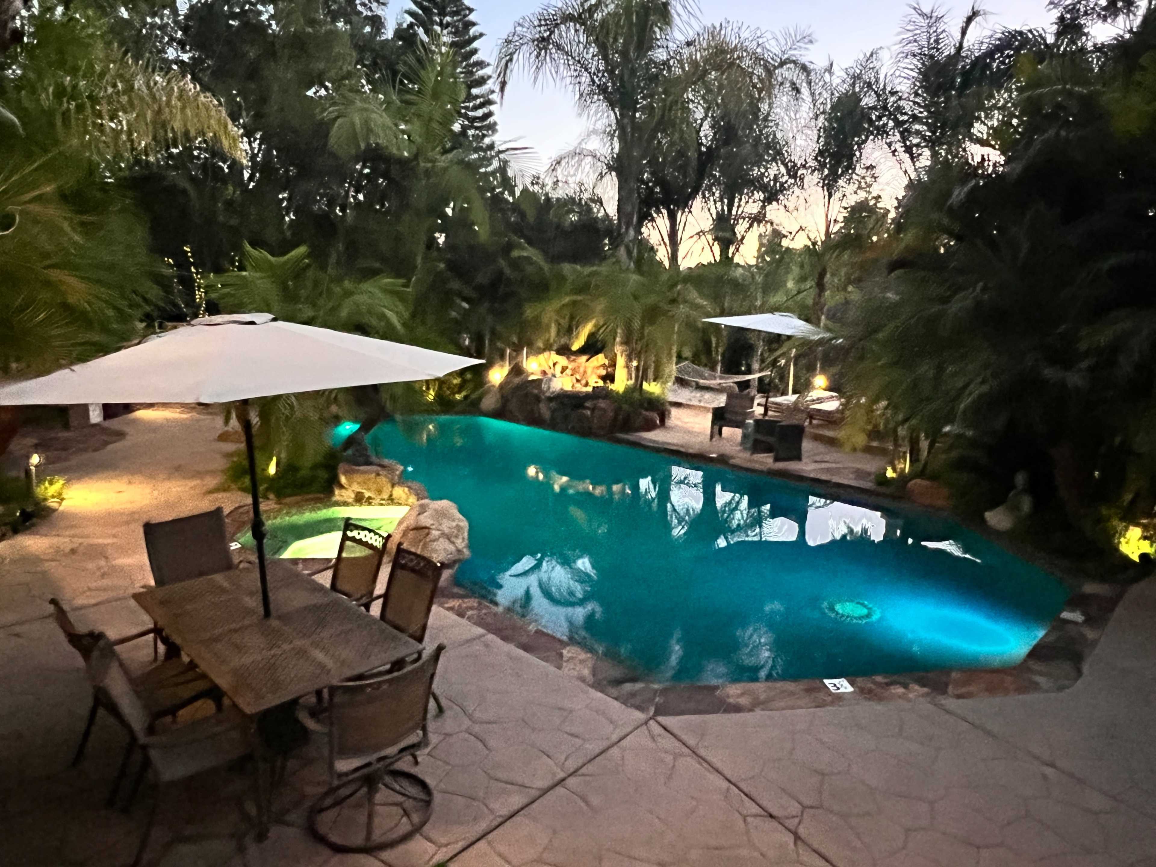 A well-lit swimming pool is surrounded by lush greenery and patio furniture in a tranquil outdoor setting.