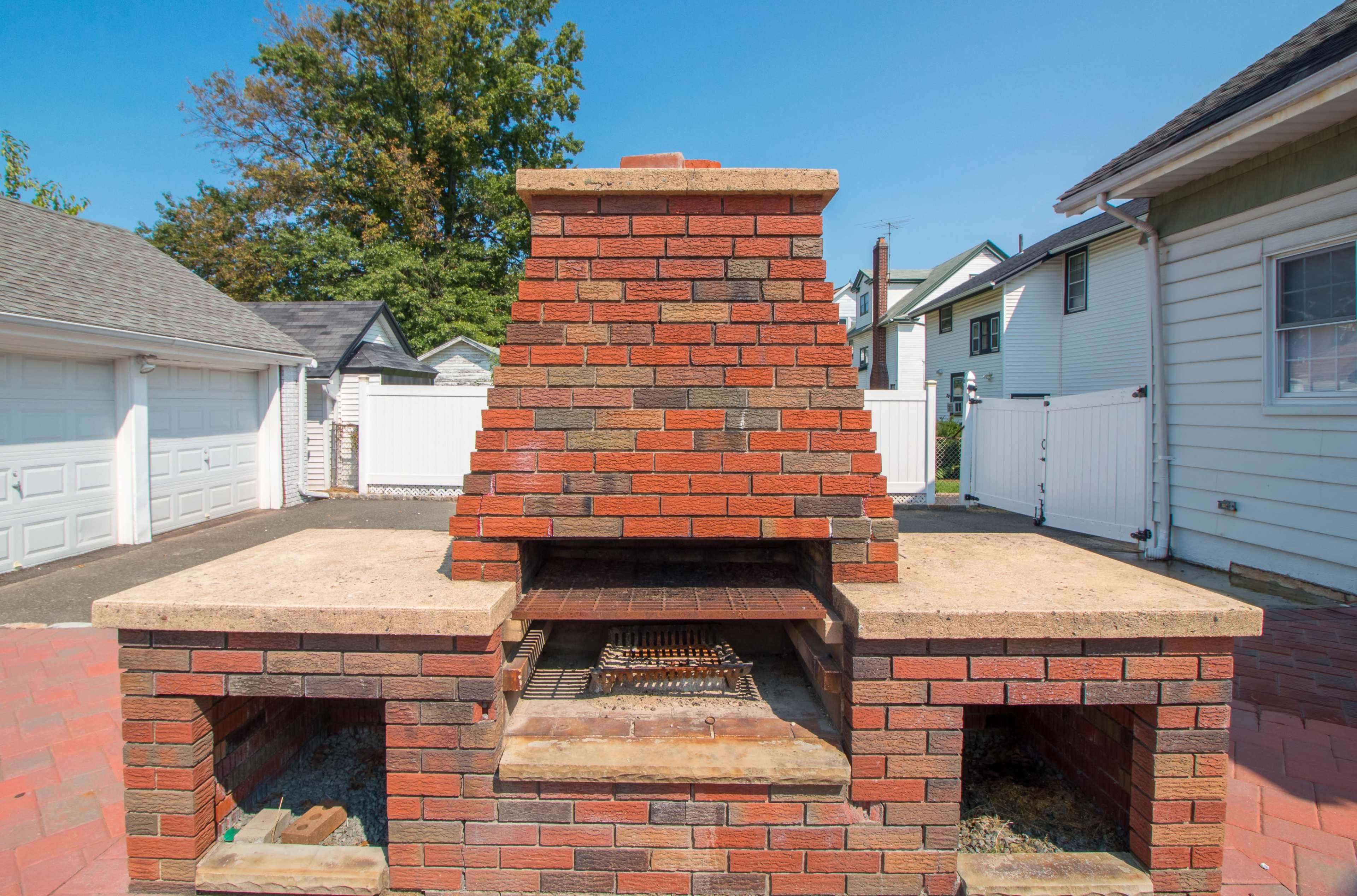A brick outdoor barbecue grill with a chimney, surrounded by a paved patio and a white picket fence.