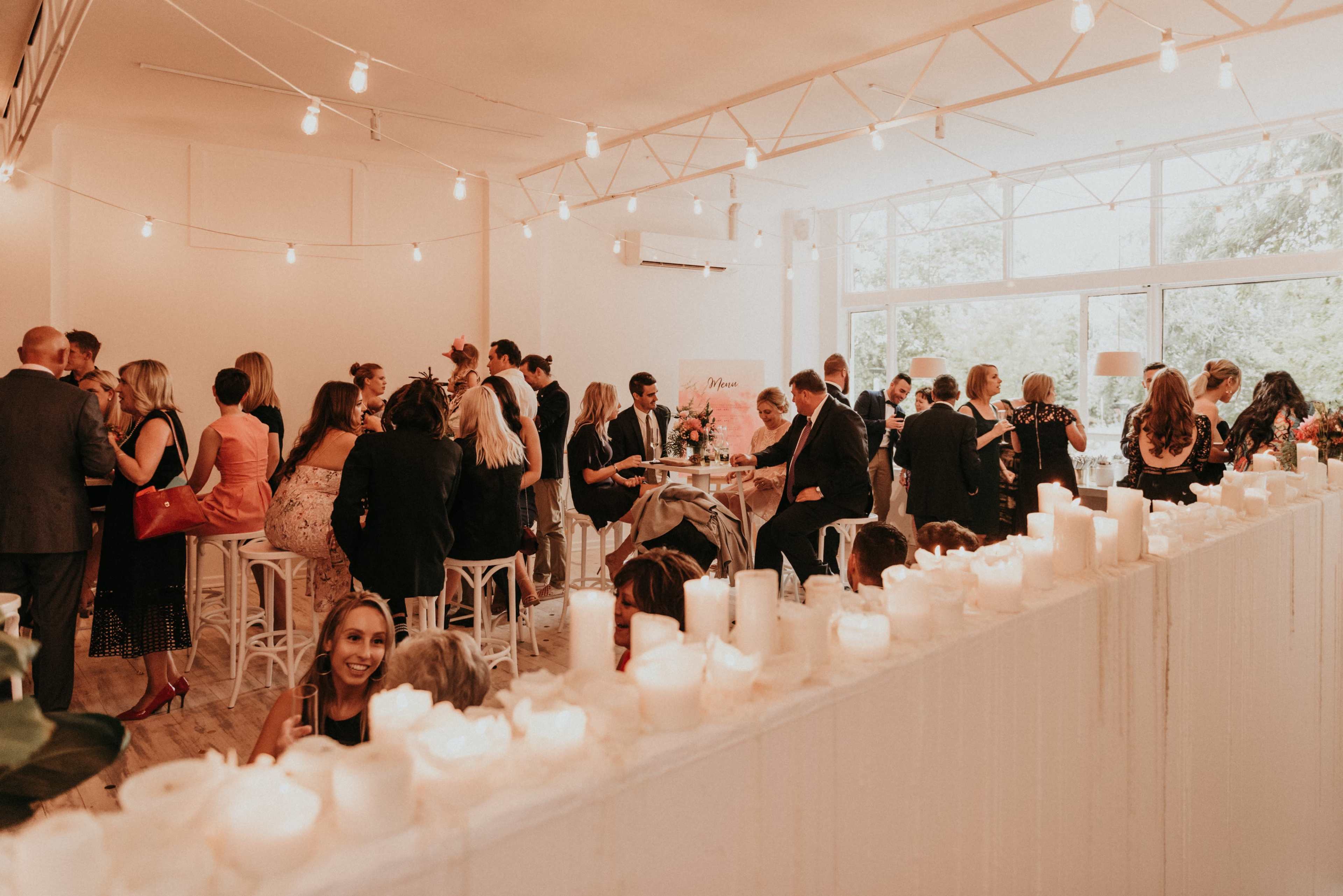 A lively gathering takes place in a brightly lit, modern venue, with guests mingling and seated around tables adorned with candles.