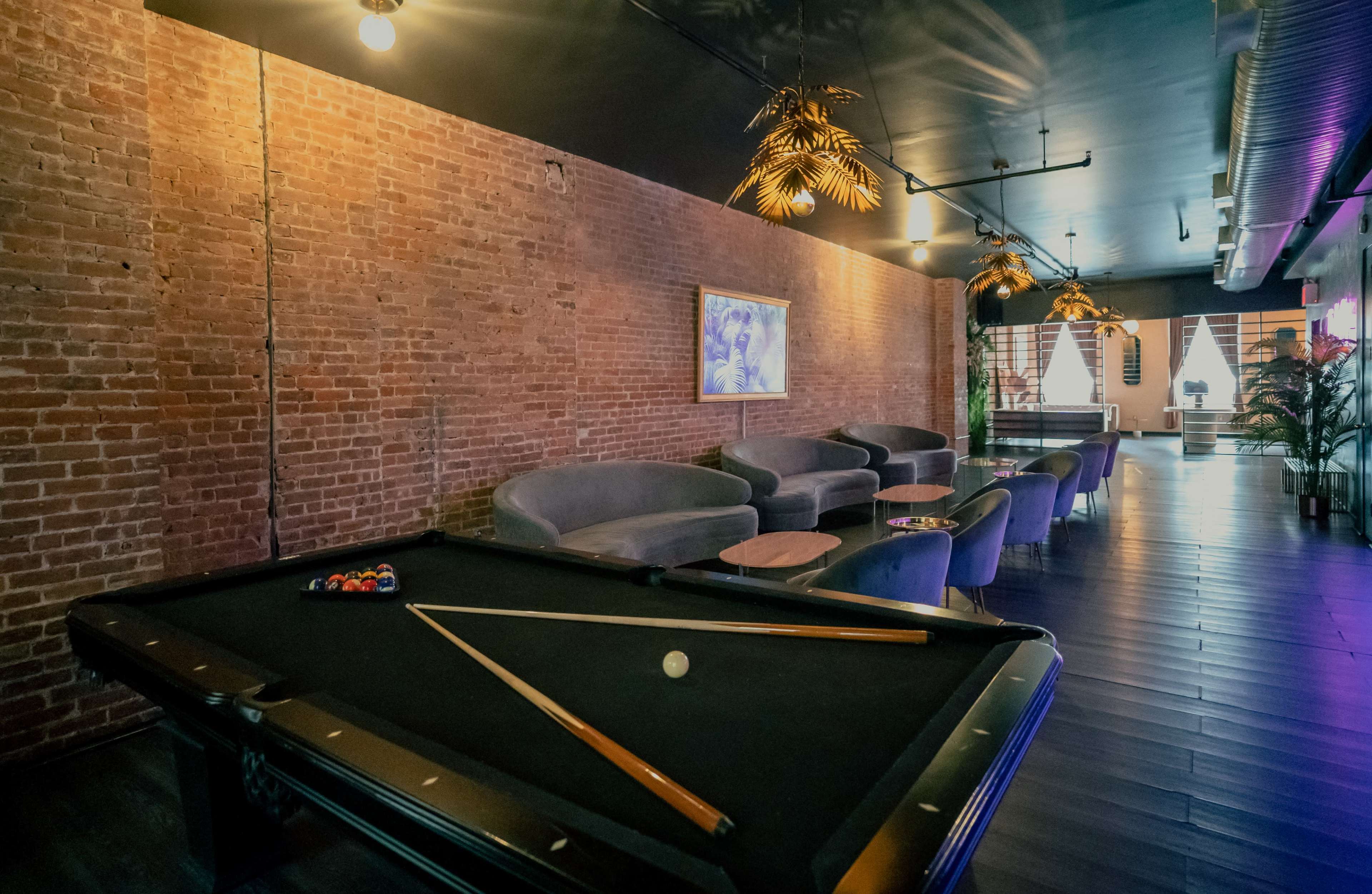 The image shows a lounge area with a billiards table in the foreground and stylish seating arrangements along a brick wall.