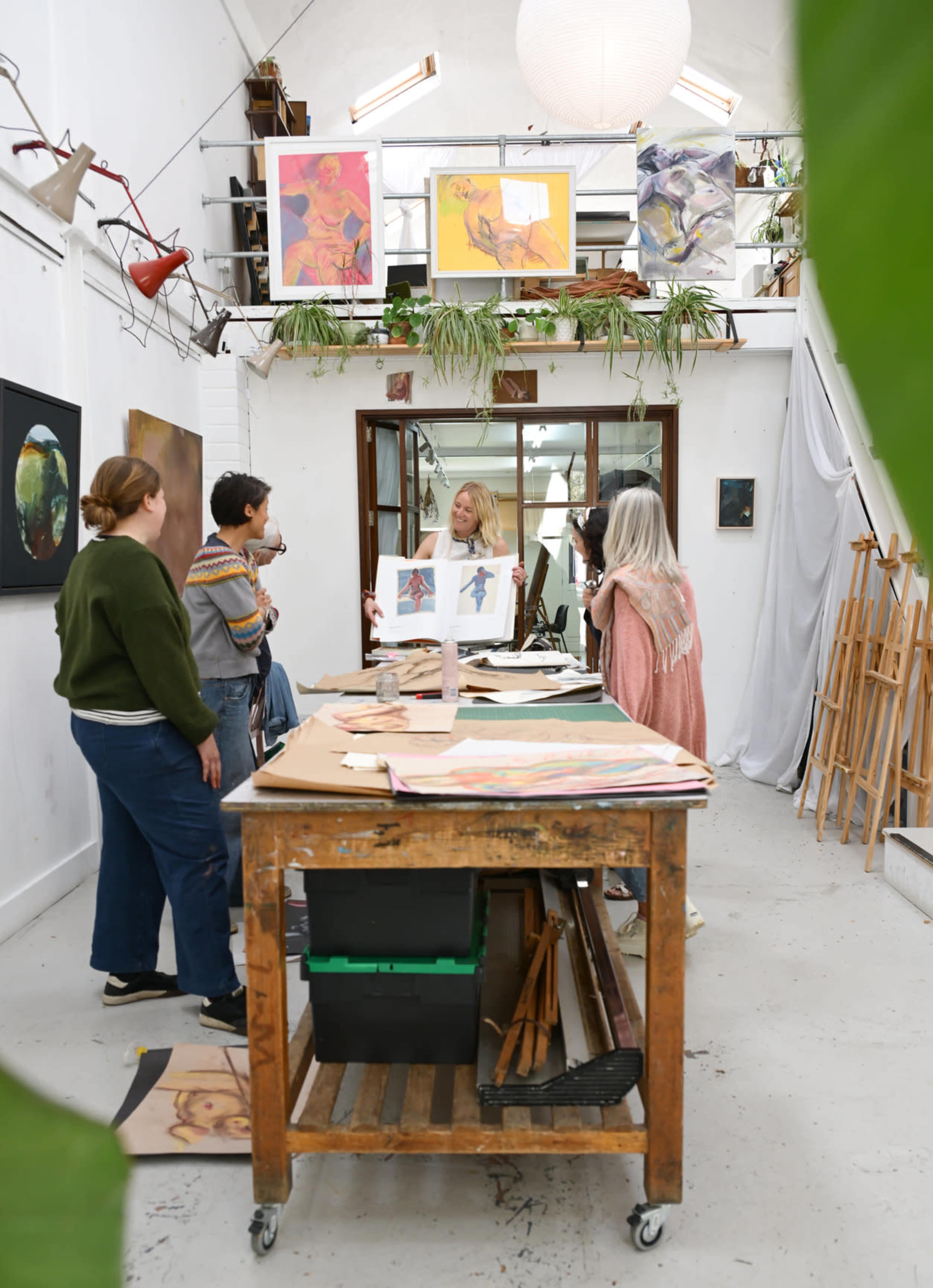 A group of individuals converse in an art studio filled with various artworks and drawing materials.