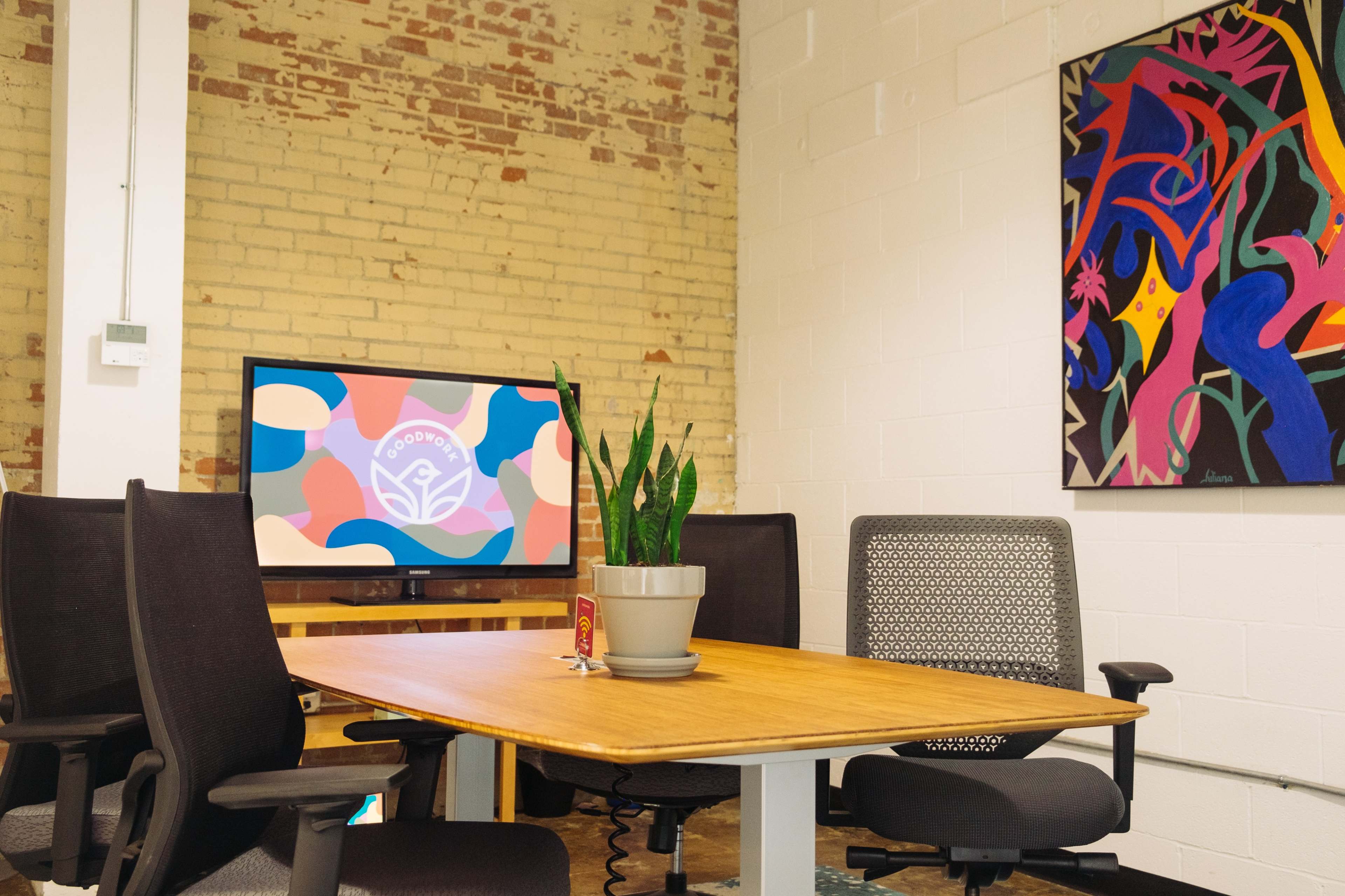 A conference room features a wooden table, four chairs, a potted plant, and a wall-mounted screen displaying a colorful graphic, with a vibrant artwork on an adjacent wall.