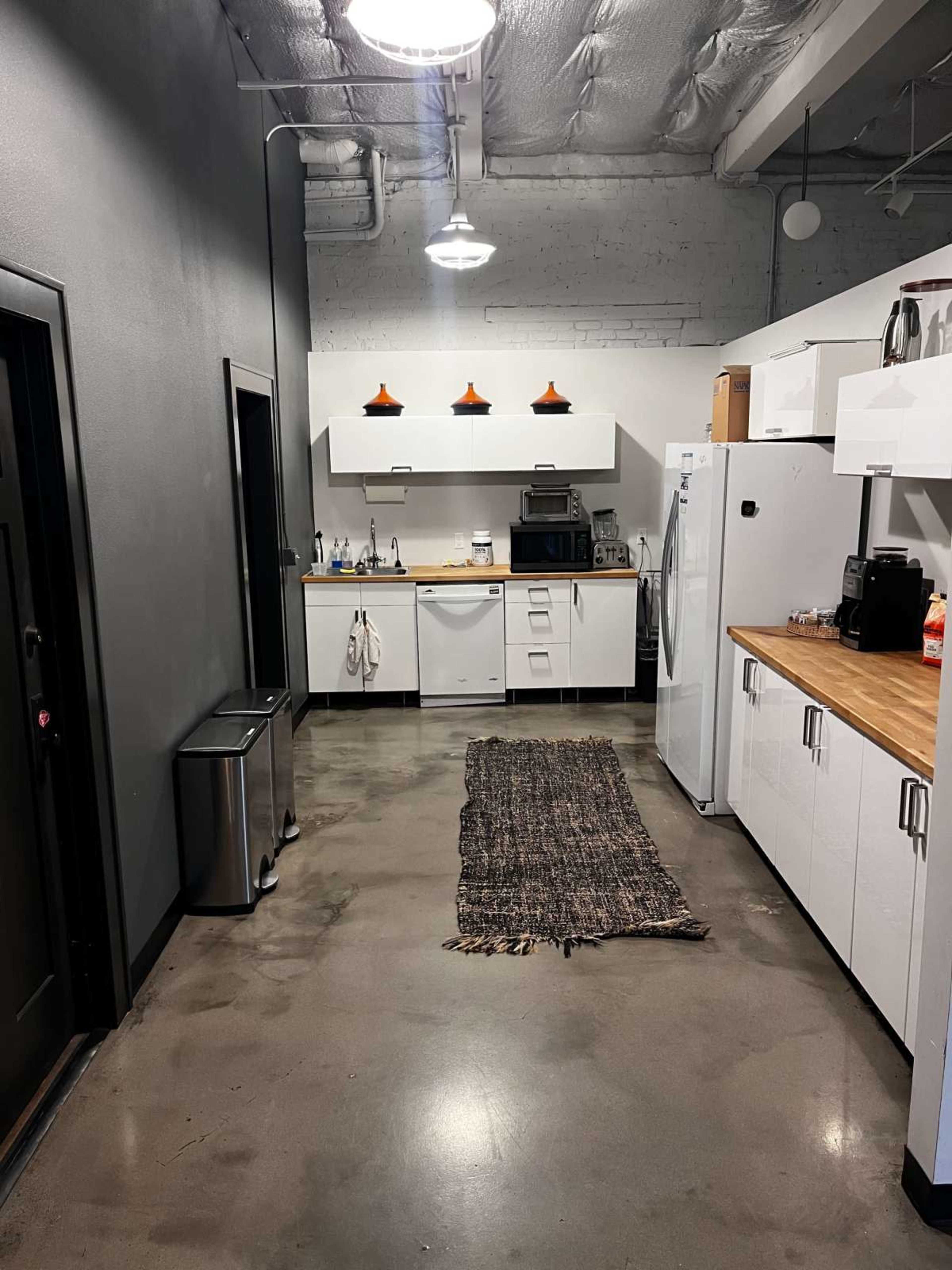 The image shows a modern, minimalistic kitchen with white cabinets, a wooden countertop, and stainless steel appliances, along with a rug running down the center of the space.