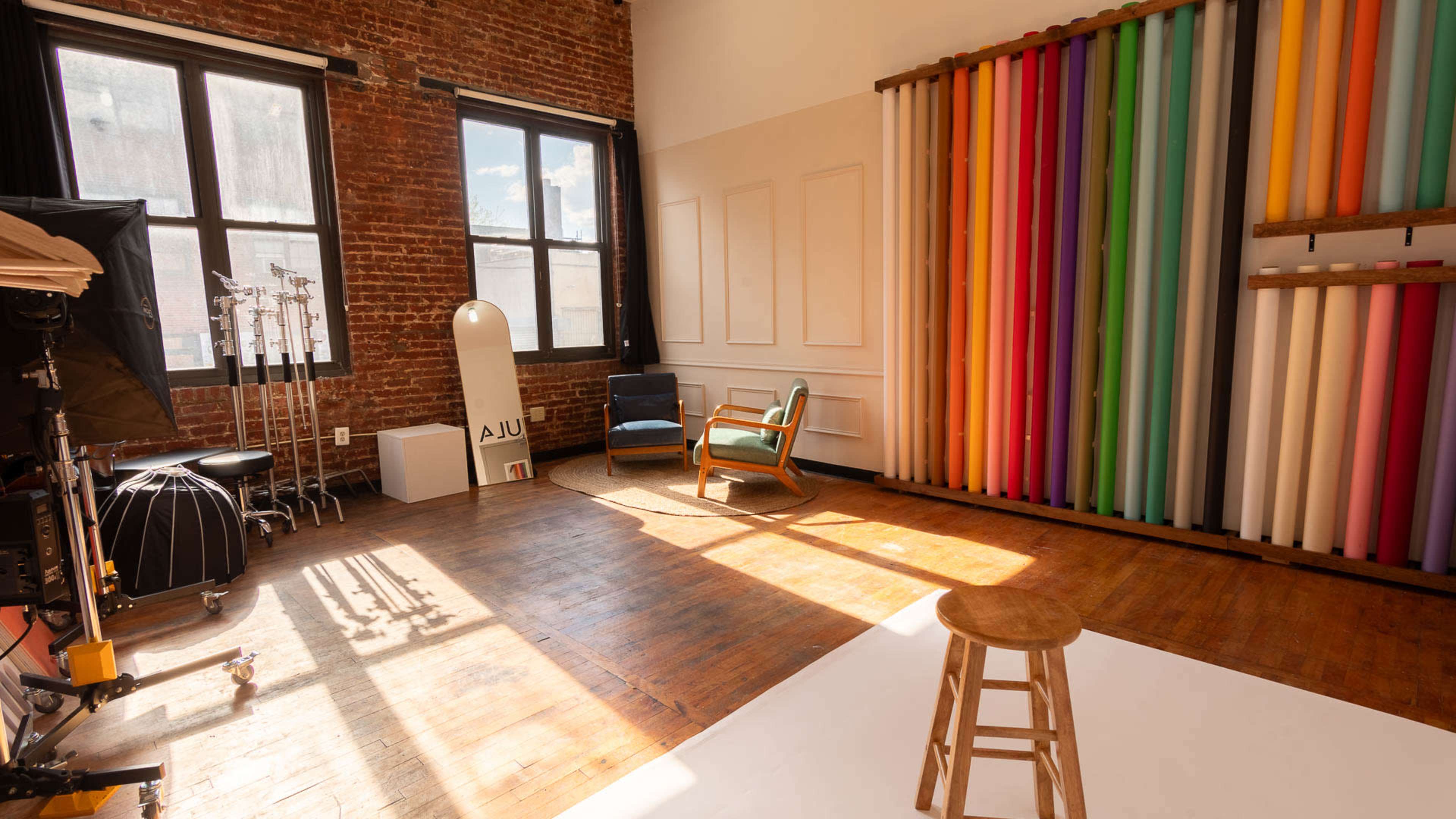 The room features a wooden floor, a stool in the center, and a wall with colorful paper rolls alongside a vintage chair and a large window letting in natural light.