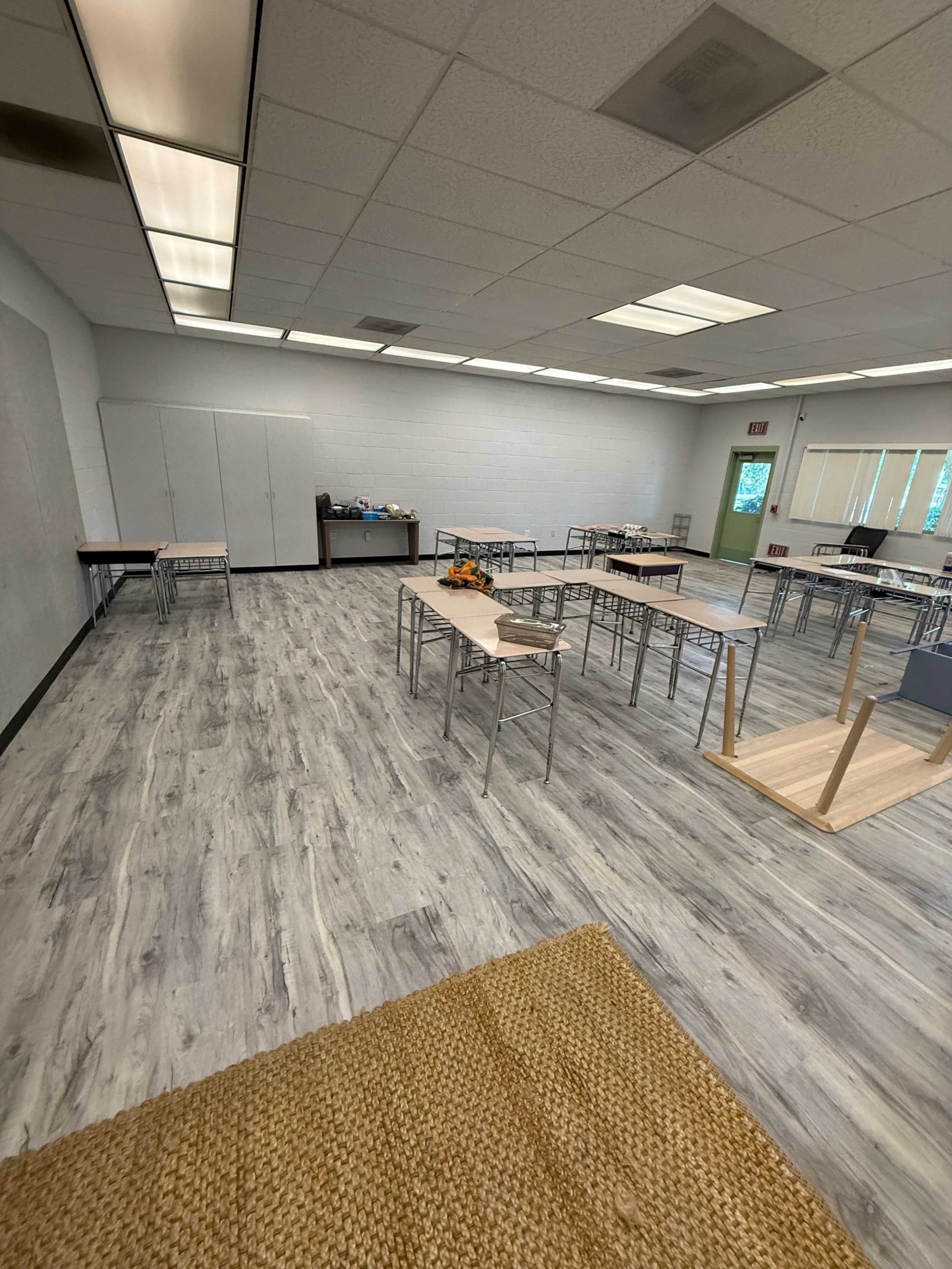 The image shows a spacious classroom with light-colored wooden flooring and several desks arranged in rows.