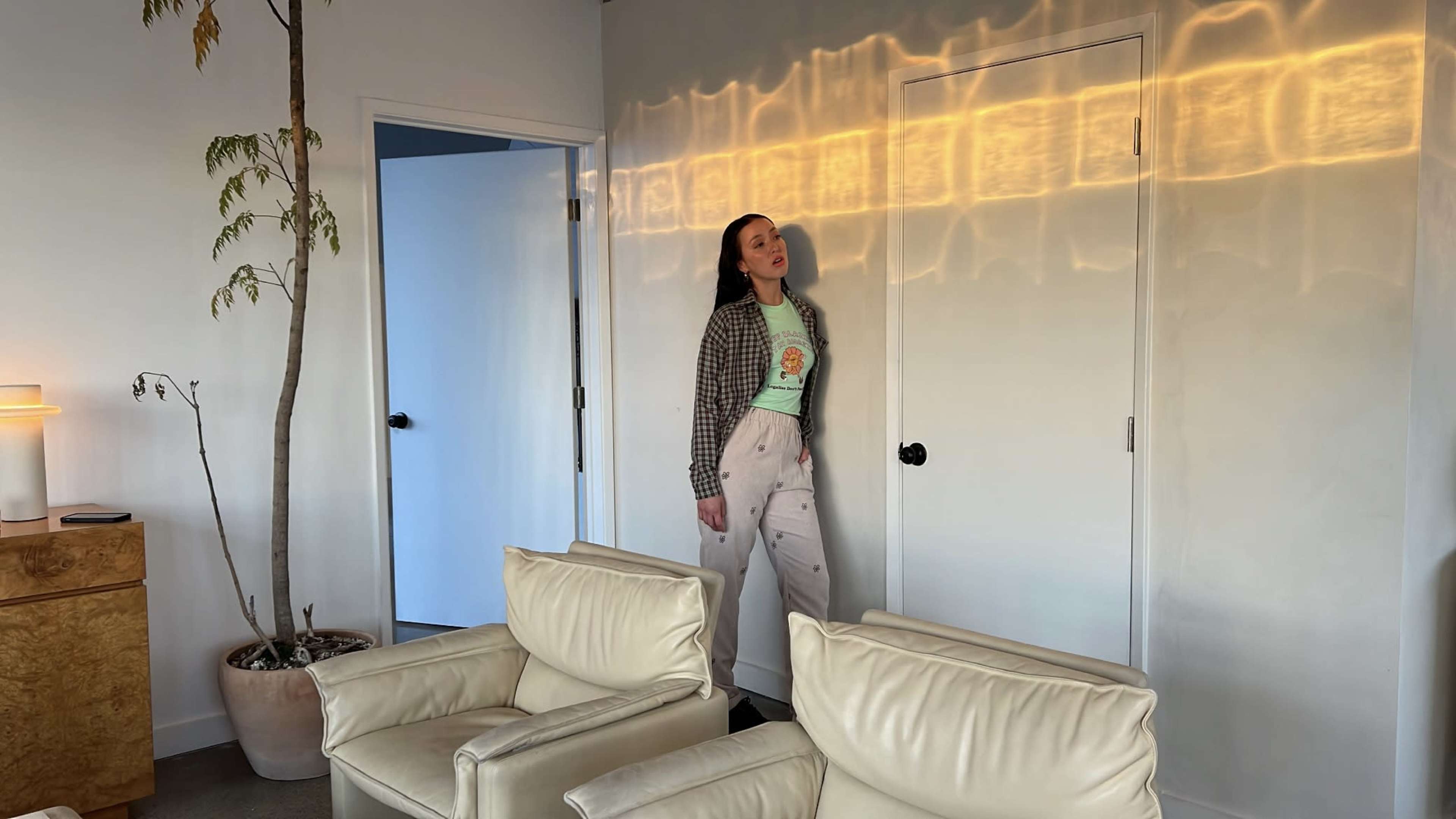 A person stands near a closed door in a minimalist room with white walls, soft lighting, and light-colored furniture.