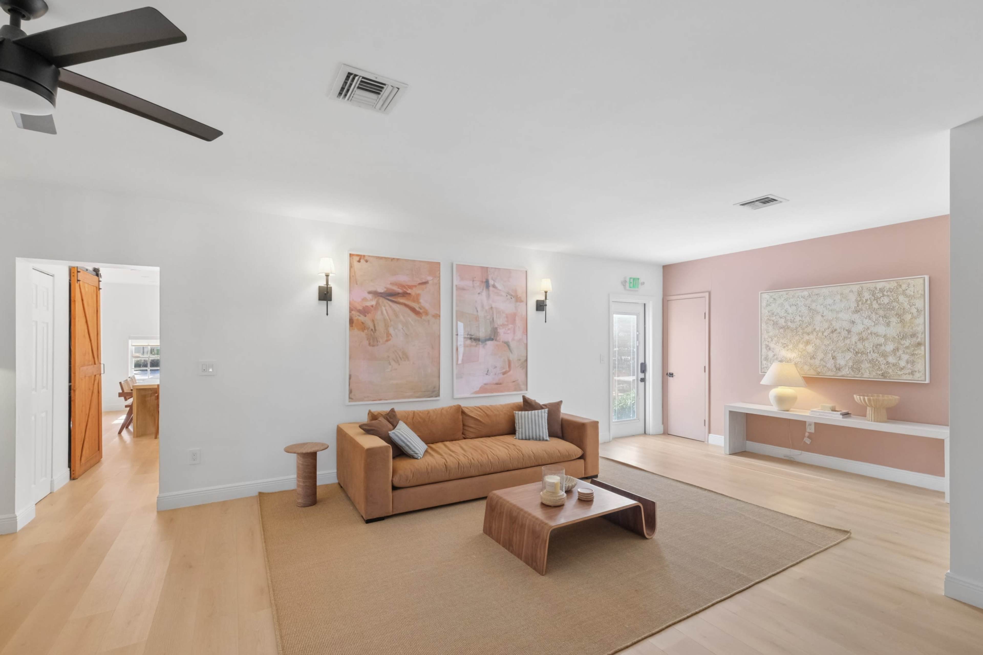 The image shows a modern living room with a tan sofa, two large abstract art pieces on the wall, a light wood coffee table, and an entryway leading to another room.