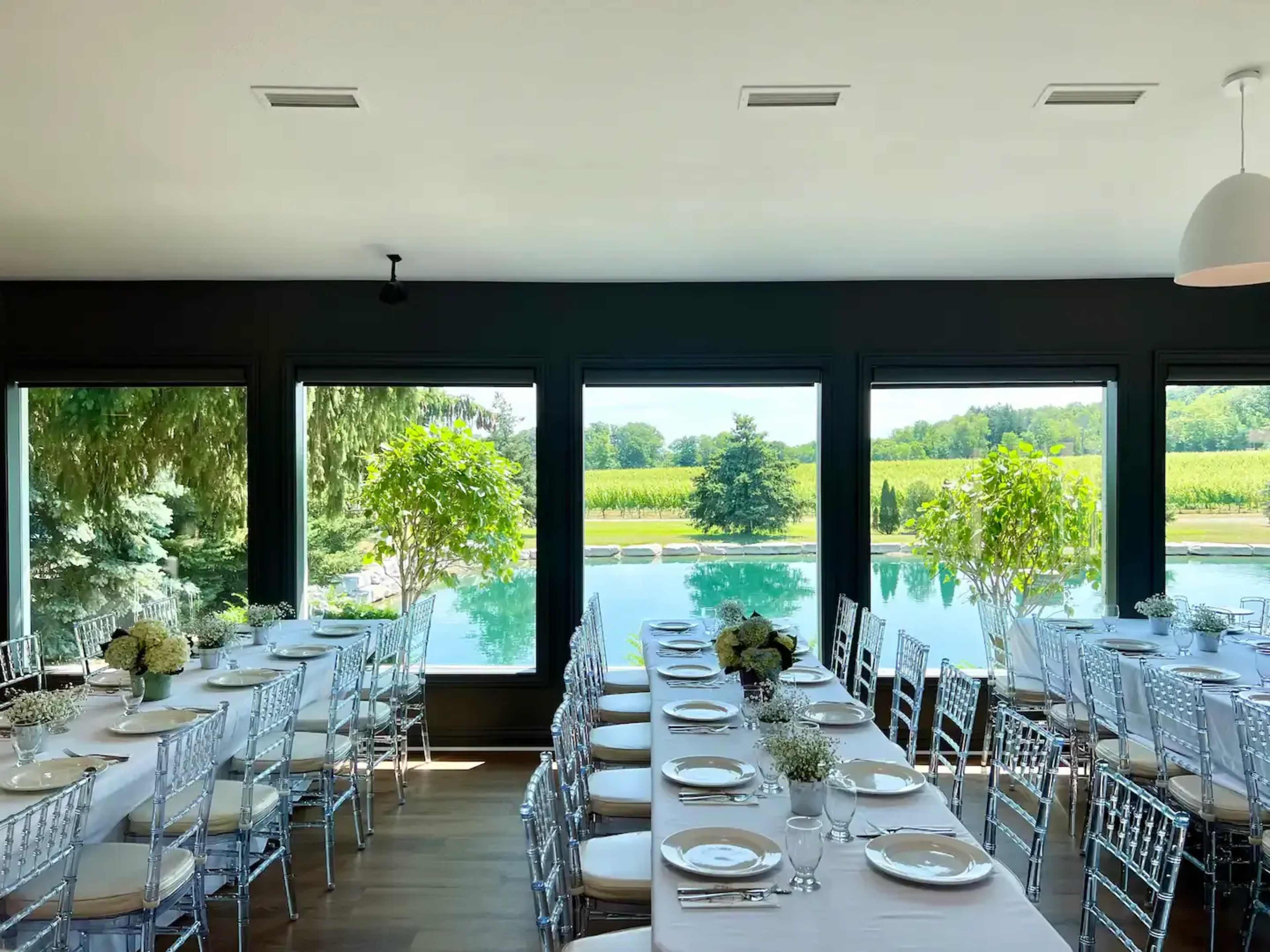 A banquet room with long tables set for a meal overlooks a serene body of water and lush greenery through large windows.