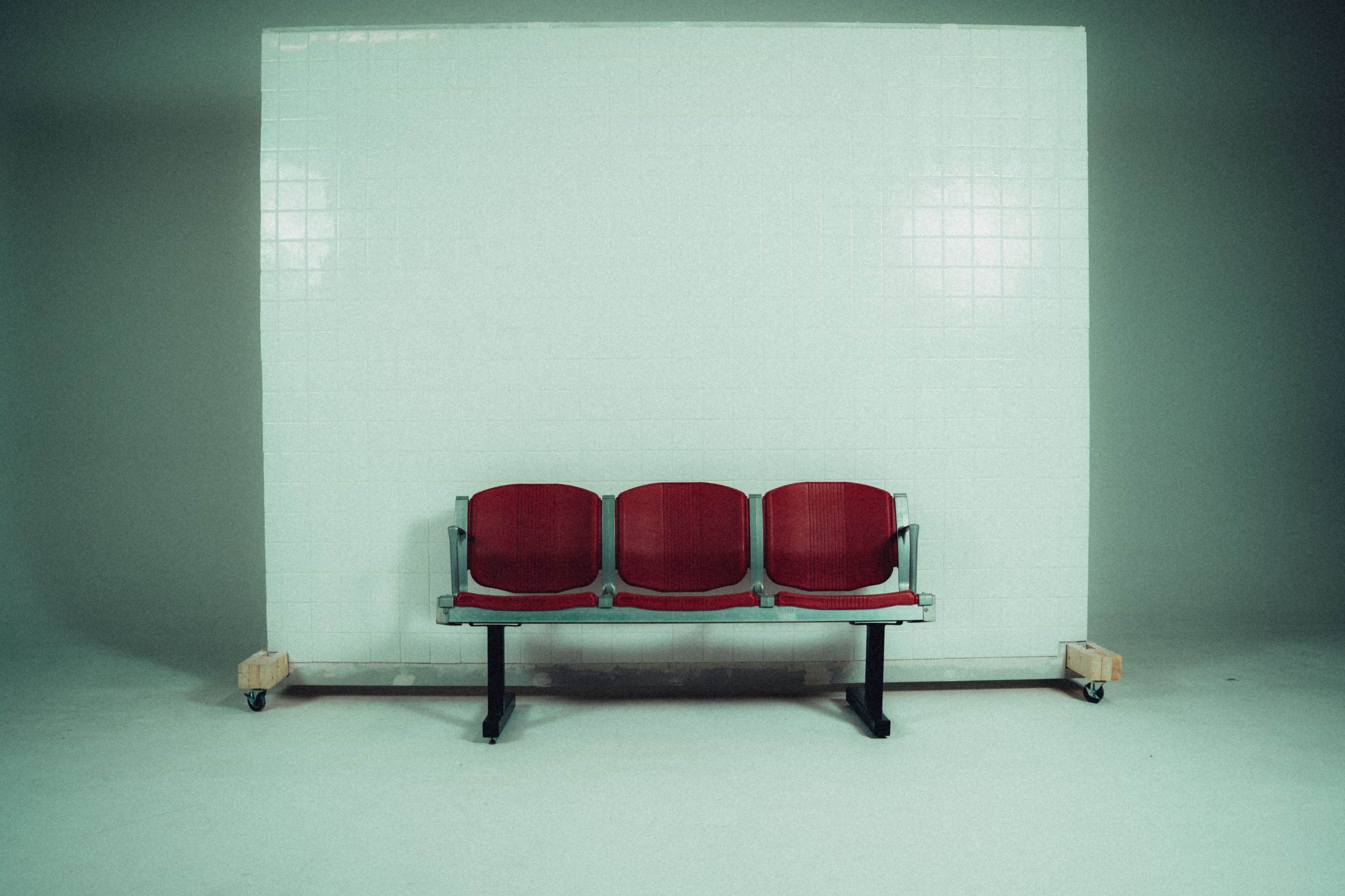 A red three-seater bench is positioned in front of a plain white tiled wall.