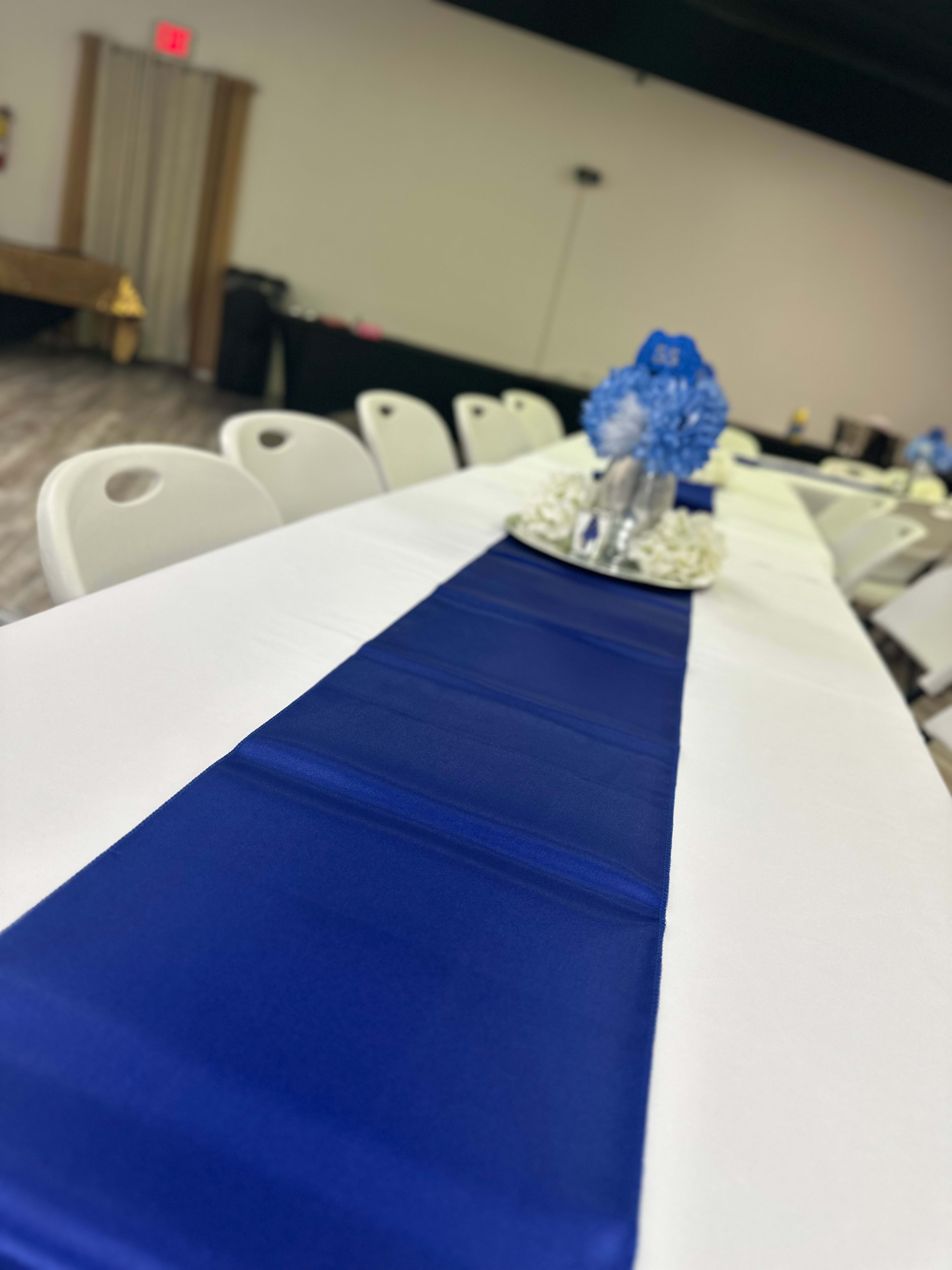A long table is set with a white tablecloth and a blue runner, featuring a centerpiece of flowers in a vase.