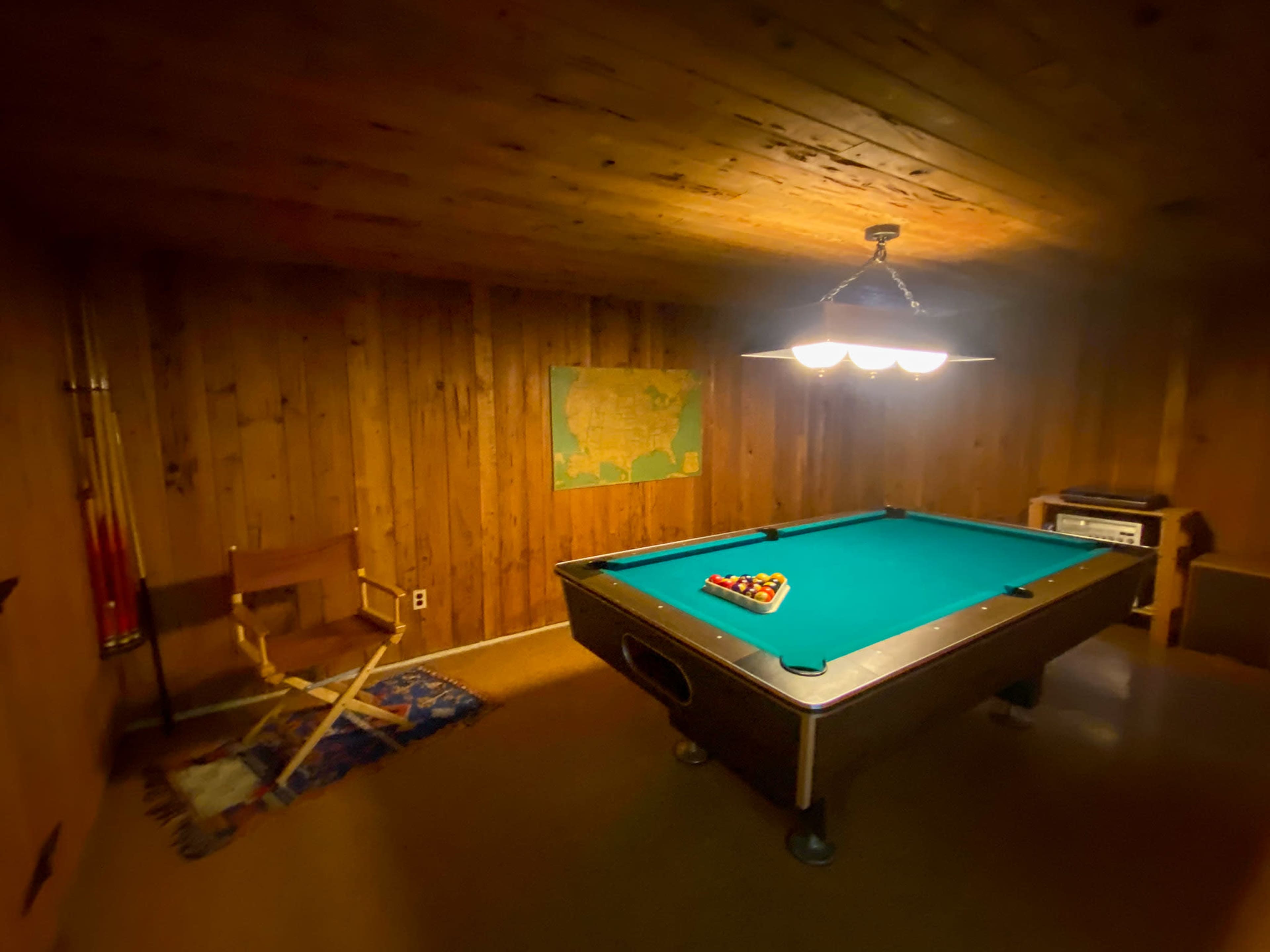 The image shows a pool table with a triangle of balls arranged on it in a dimly lit room decorated with wooden walls and a map on the corner.