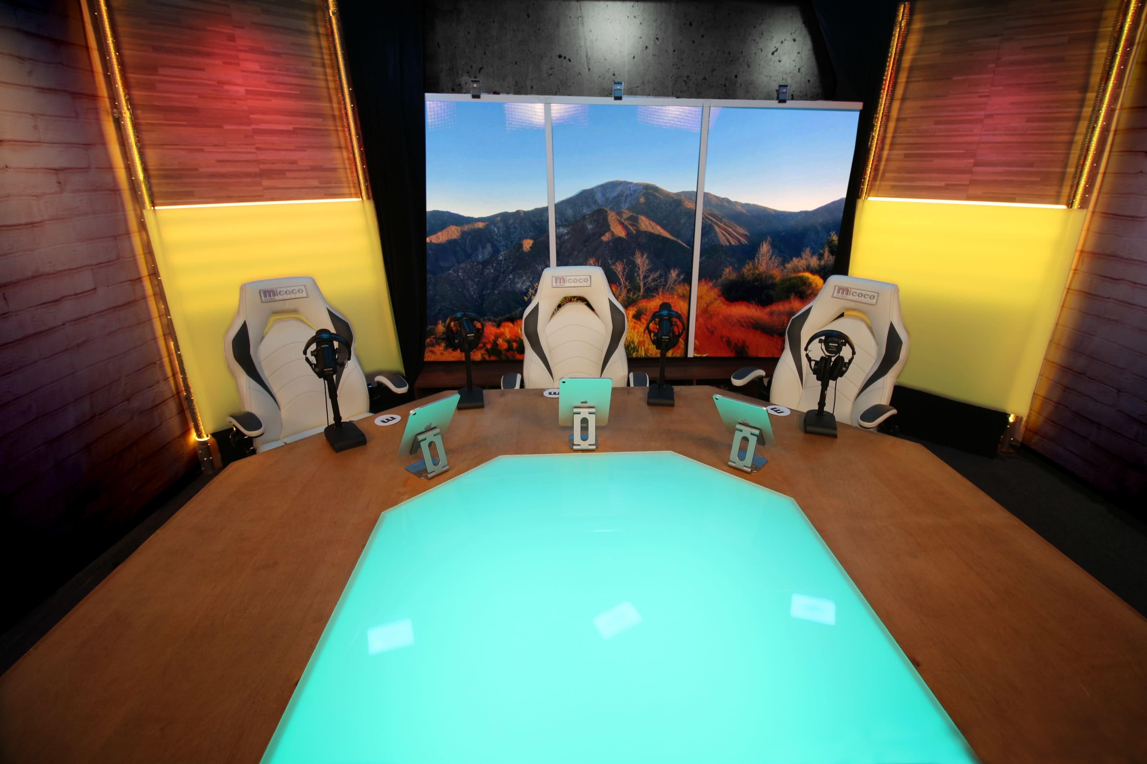 The image shows a modern studio setup featuring three high-backed chairs arranged around a hexagonal table with a glowing surface, illuminated by colorful lights and a backdrop of mountains displayed on screens.