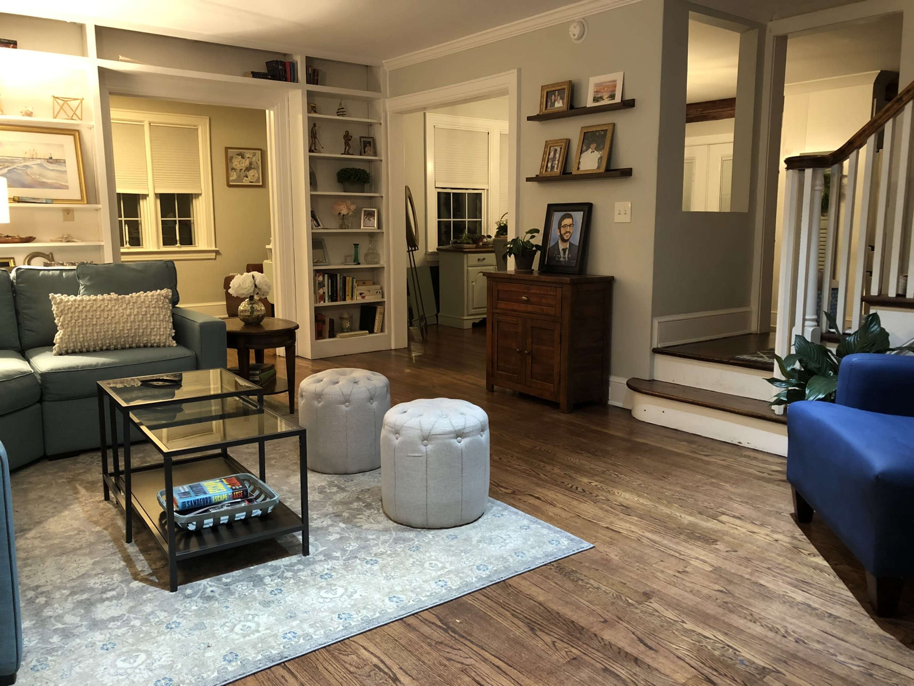 A cozy living room featuring a blue sofa, a glass coffee table, two ottomans, and a light wooden floor.