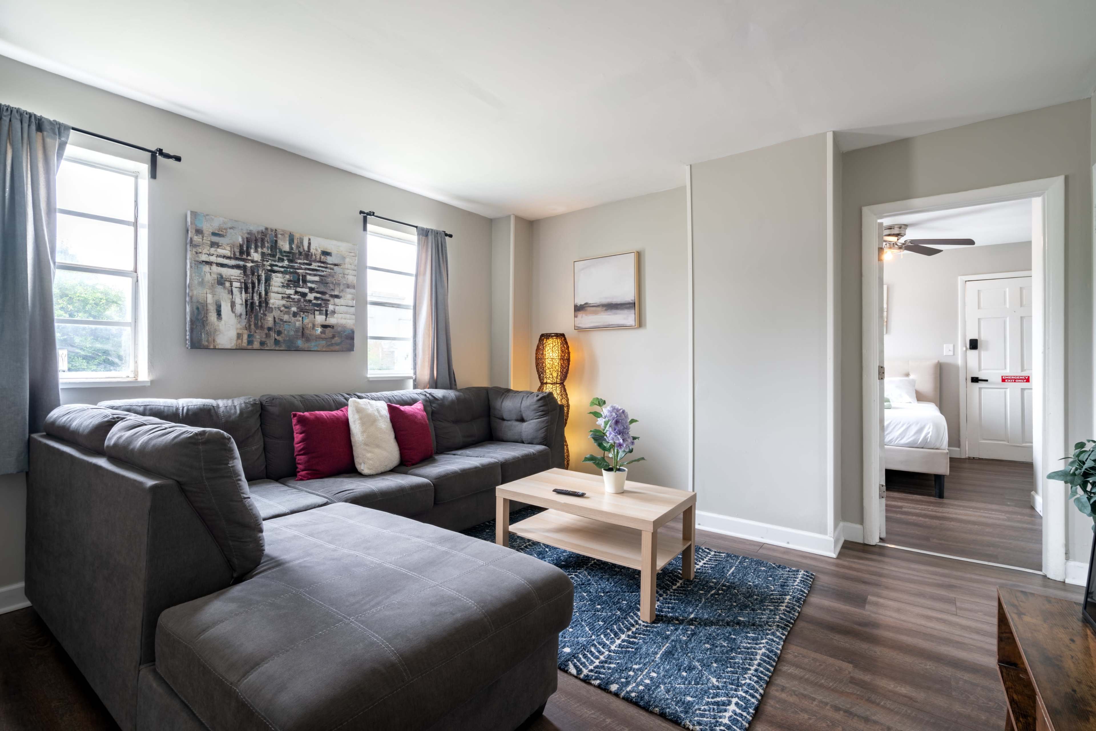 The image shows a modern living room with a gray sectional sofa, a light wooden coffee table, and a doorway leading to a bedroom.