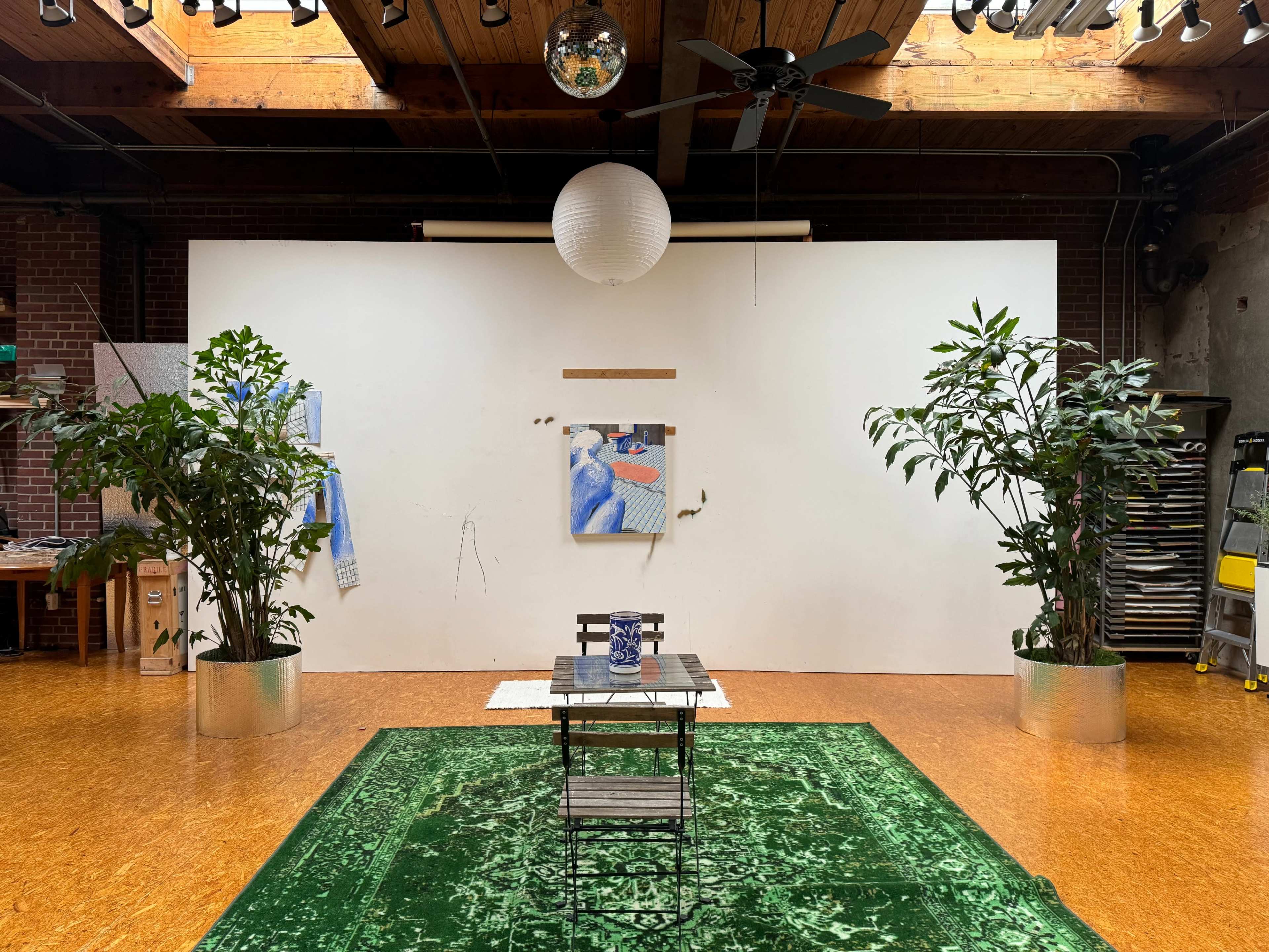 The scene features a spacious studio with a large white wall adorned by a painting, a wooden table with chairs placed on a green rug, and greenery in metallic planters.