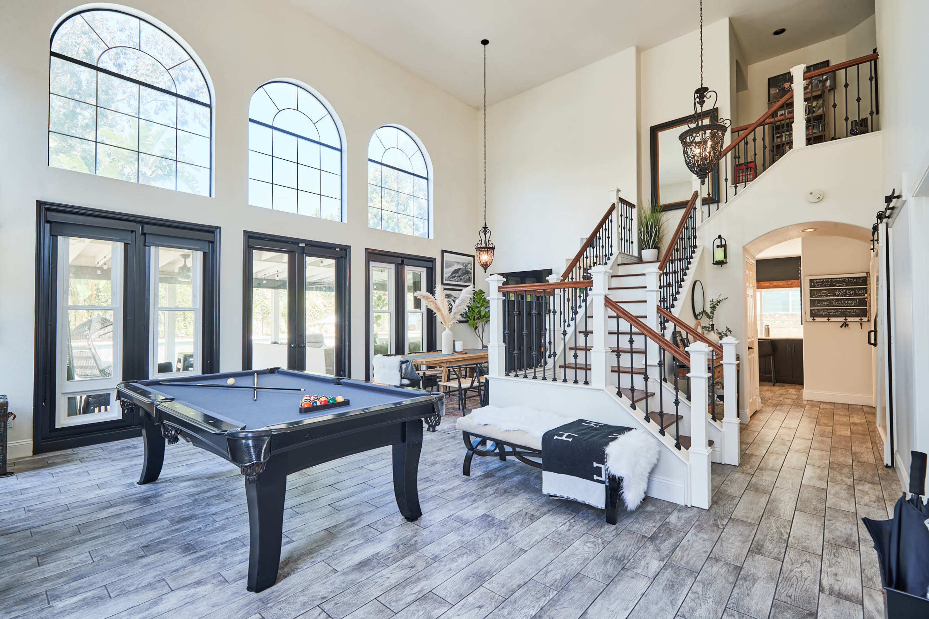 A spacious, well-lit room featuring a billiard table, large arched windows, and a staircase leading to an upper level.