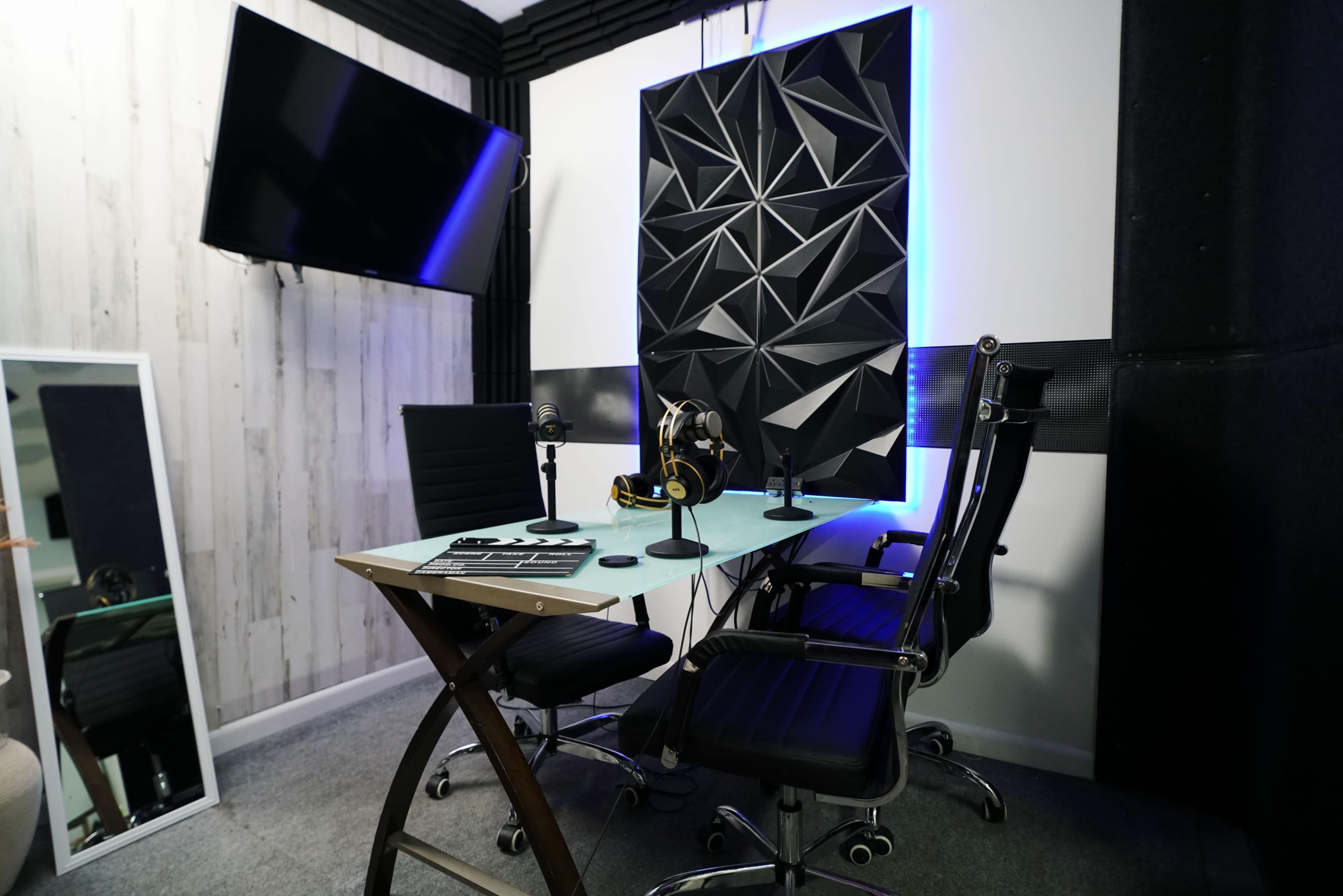 A recording studio with a glass table, two black ergonomic chairs, microphones, and soundproof wall panels.