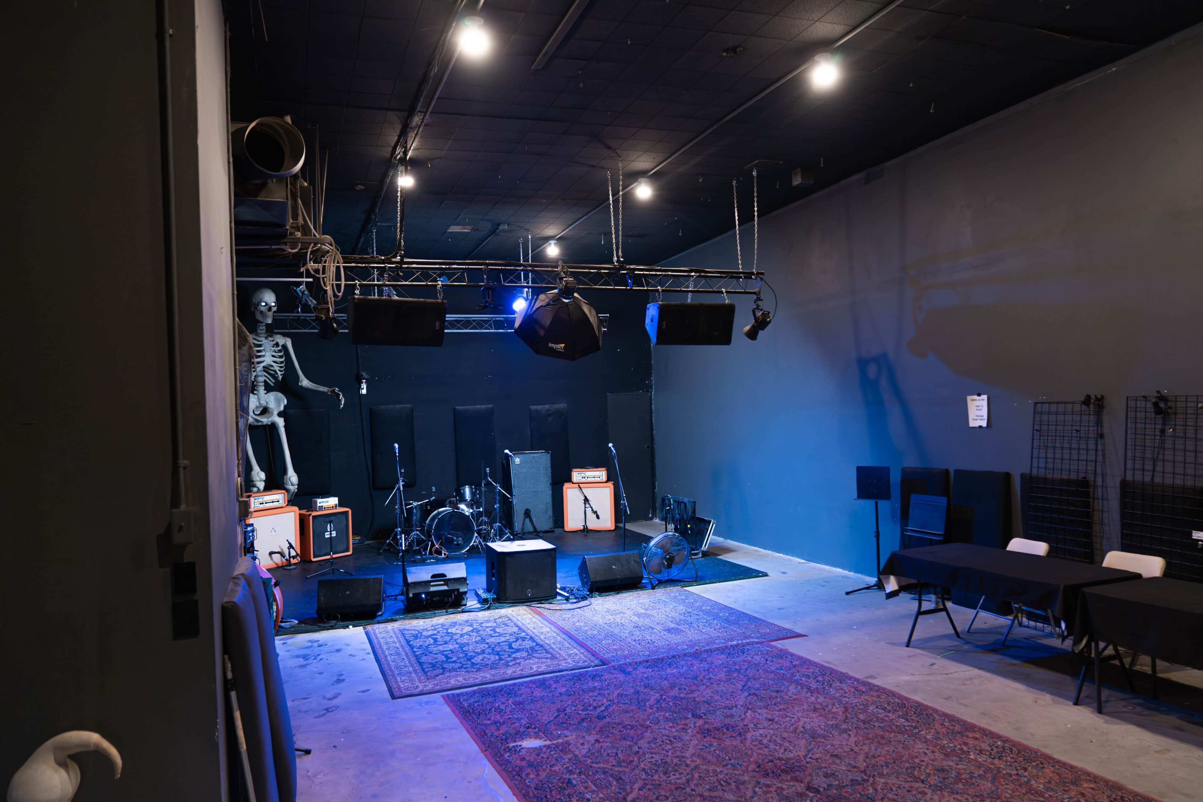 The image shows a dimly lit music venue featuring a stage with amplifiers, a drum set, and a skeleton decoration, along with tables arranged in front.