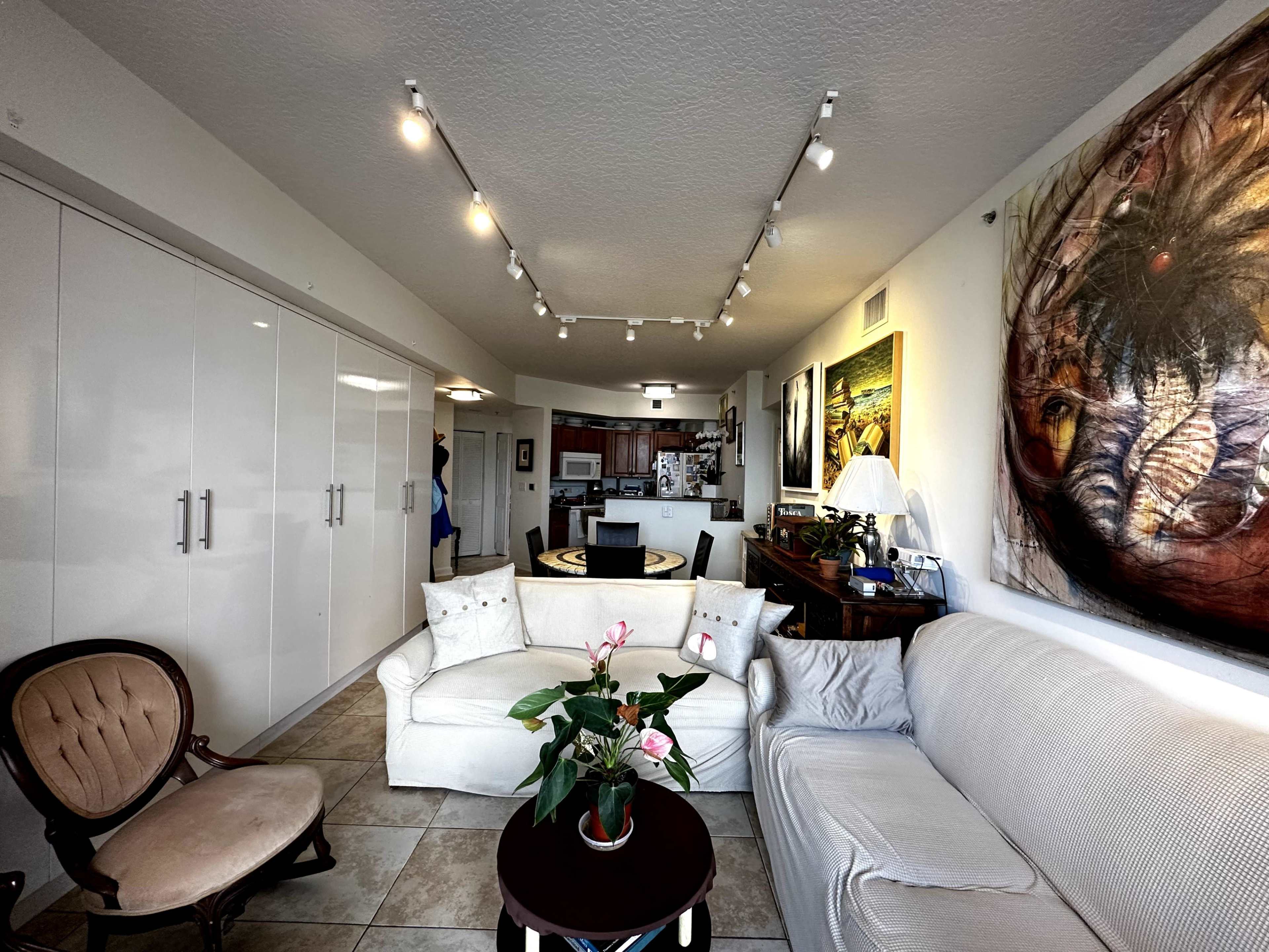 A modern living room with a white sofa, an armchair, and a dining area visible in the background, along with wall art and cabinetry.