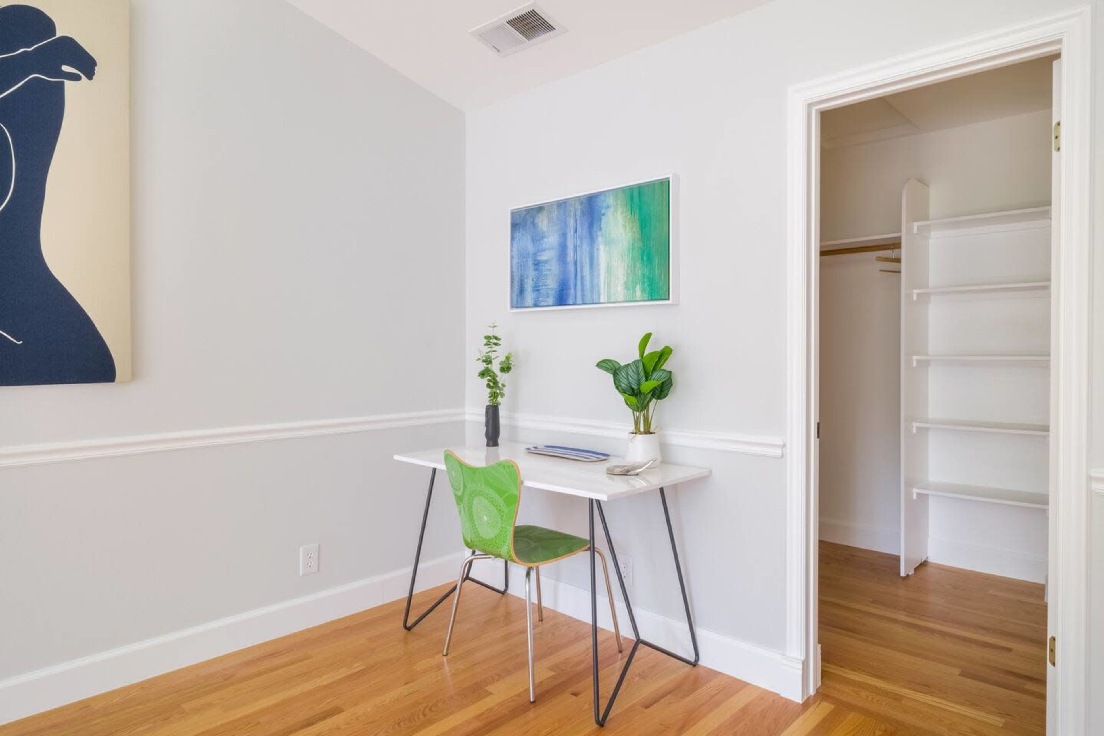 A small, well-lit office space features a minimalist desk with a green chair, a plant, and abstract artwork on the wall, alongside a closet with white shelves.